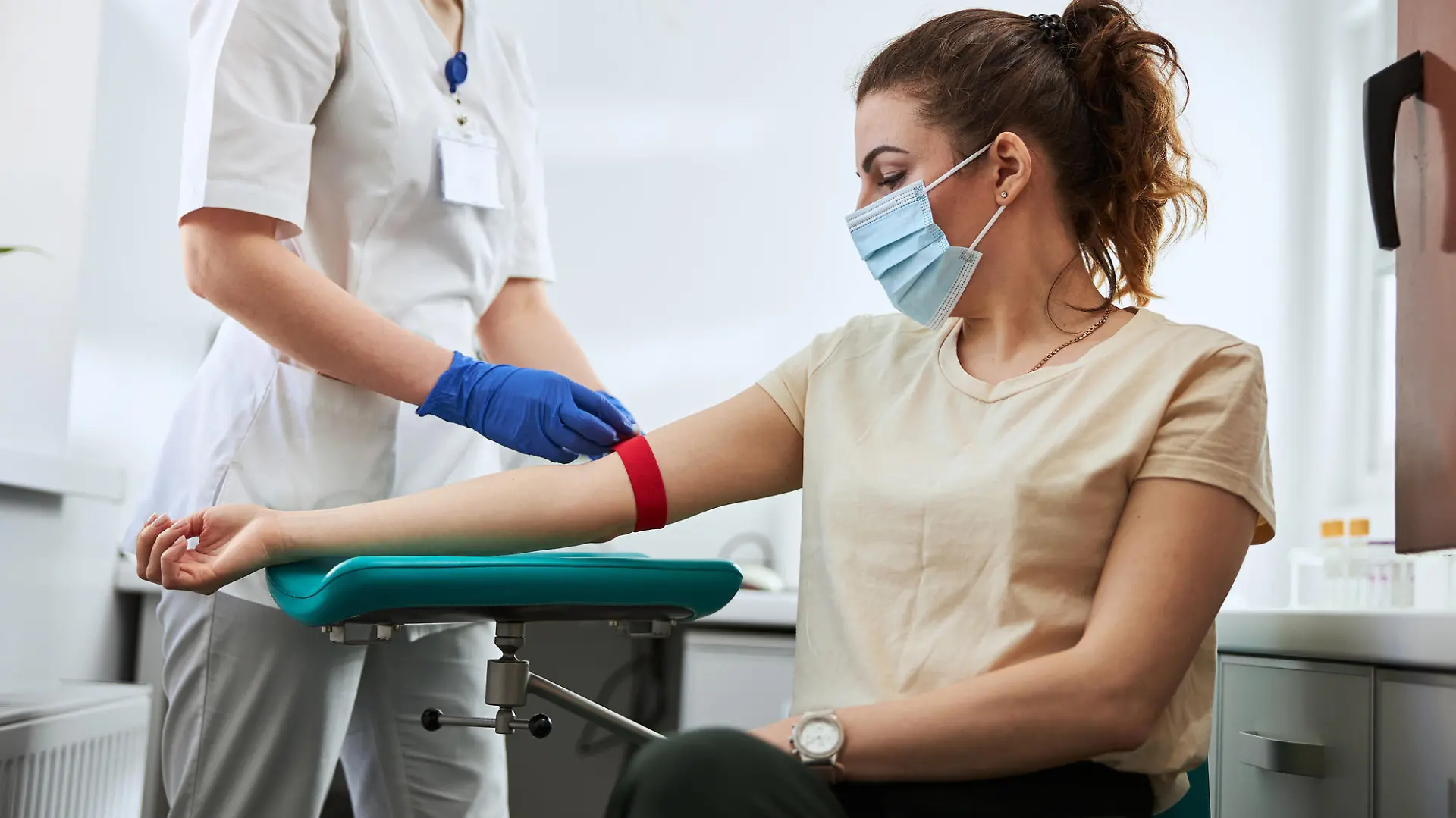 Skilled focused young phlebotomy technician applying a tourniquet to a female patient arm for venipuncture