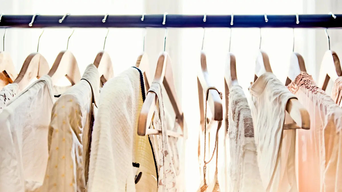 A row of light-colored clothing secured on wooden hangers is visible from about armpit level up, dangling from a rod which stretches from one end of the frame to the other. Behind the rack, two large windows brighten the room with daylight.