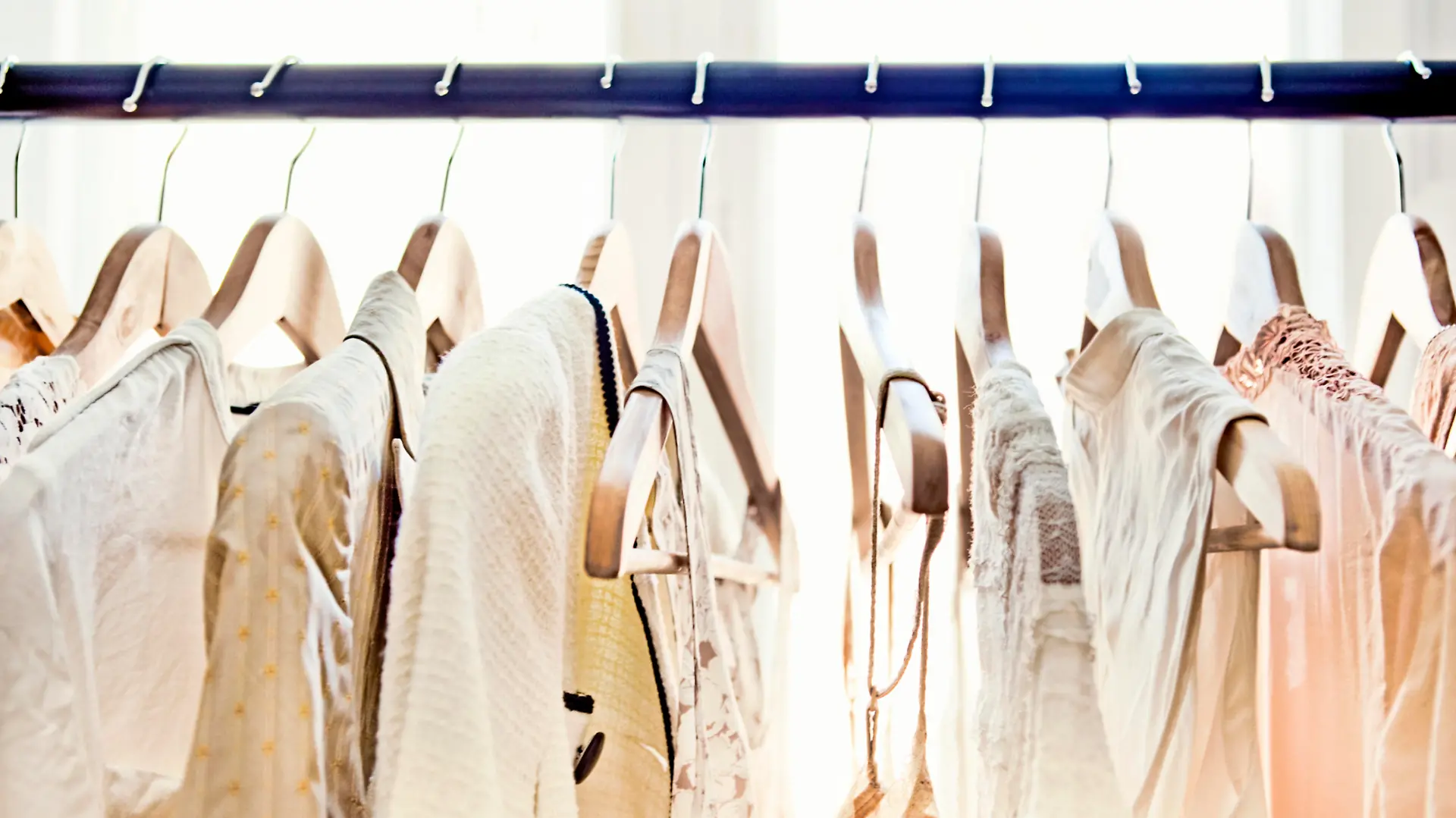 A row of light-colored clothing secured on wooden hangers is visible from about armpit level up, dangling from a rod which stretches from one end of the frame to the other. Behind the rack, two large windows brighten the room with daylight.