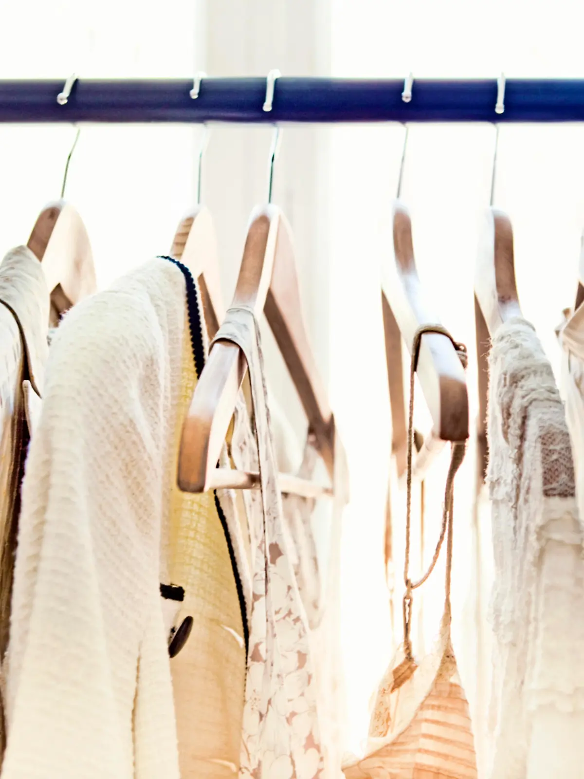 A row of light-colored clothing secured on wooden hangers is visible from about armpit level up, dangling from a rod which stretches from one end of the frame to the other.  Behind the rack, two large windows brighten the room with daylight.