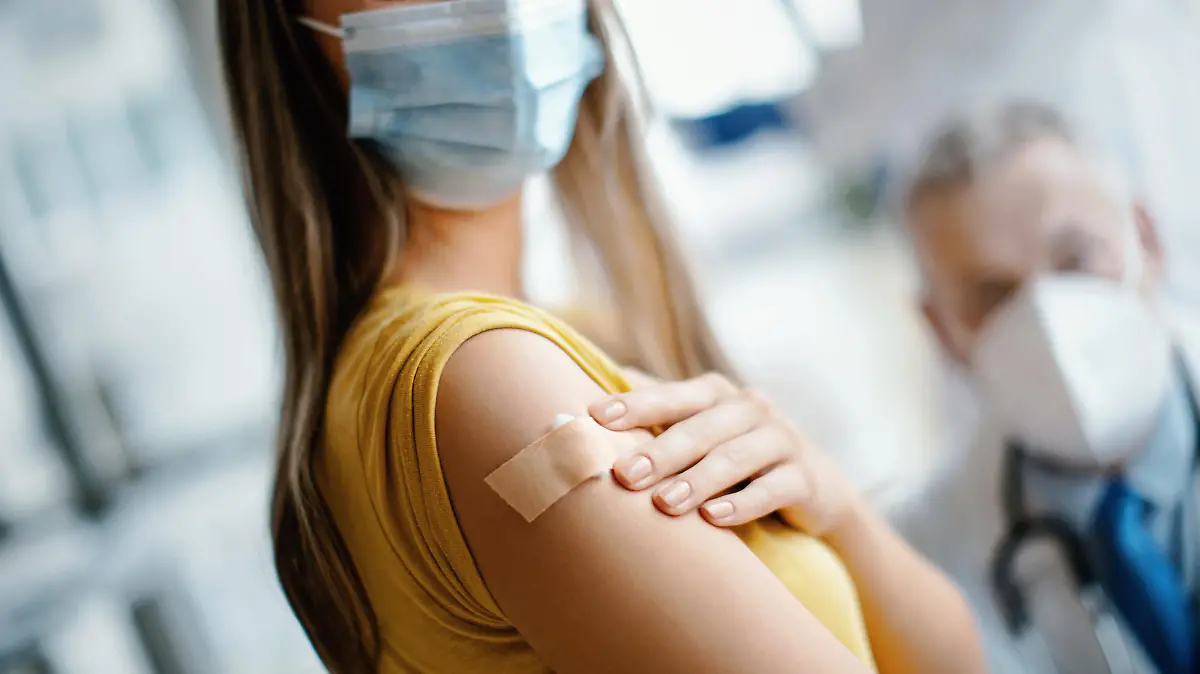 Doctor had just vaccinated a young female patient in the hospital. She is pleased with how it all went. She's smiling behind a protective face mask and looking at camera. Focus on adhesive bandage. Medical worker is in protective workwear.