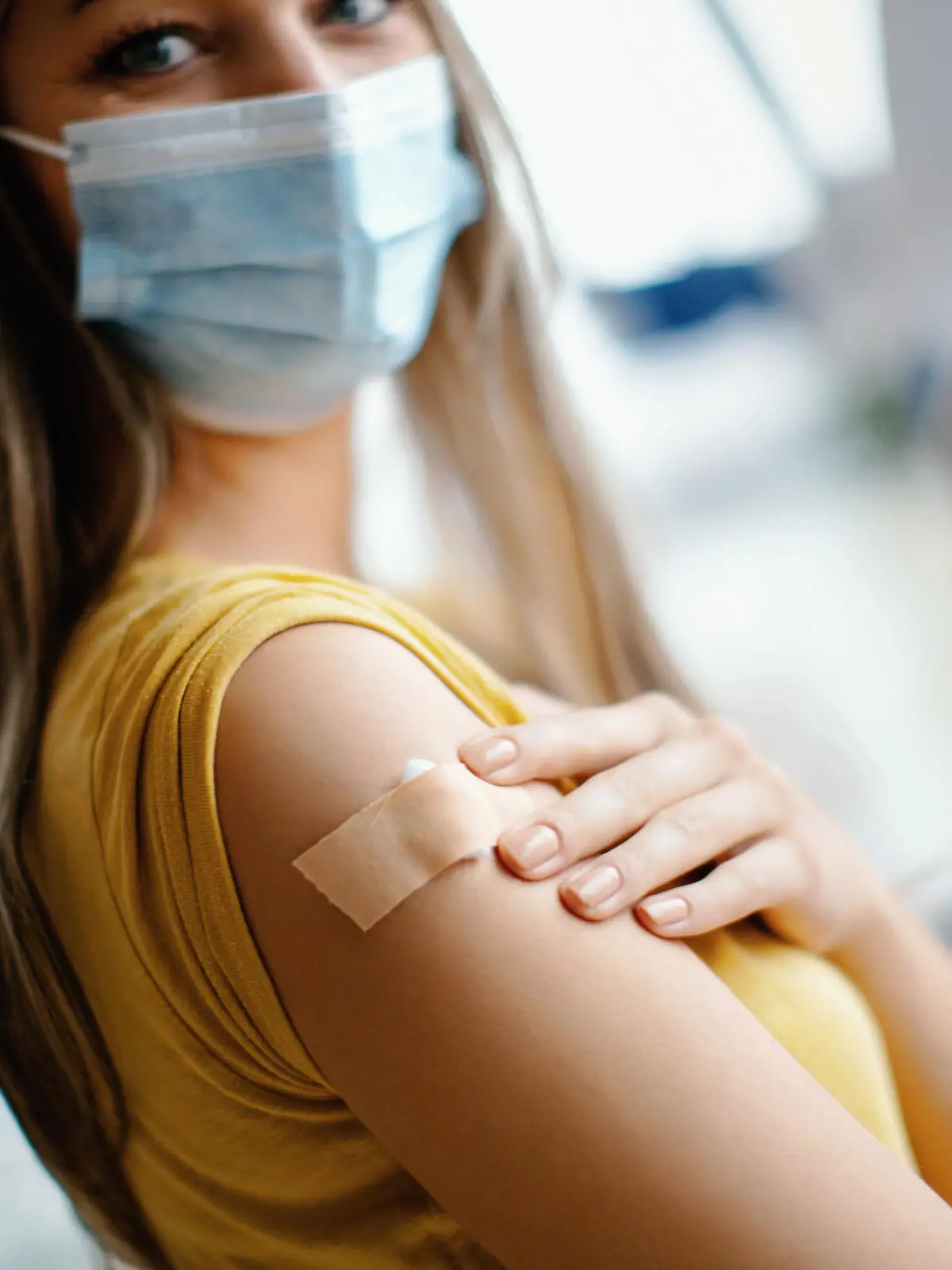 Doctor had just vaccinated a young female patient in the hospital. She is pleased with how it all went. She's smiling behind a protective face mask and looking at camera. Focus on adhesive bandage. Medical worker is in protective workwear.
