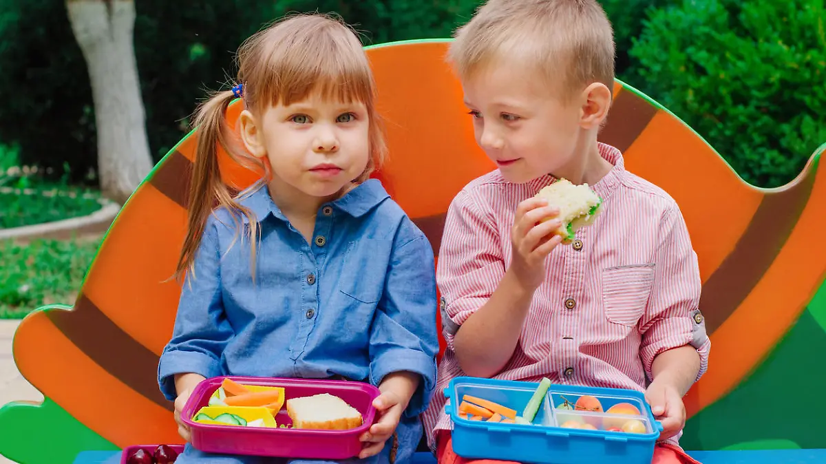 Elementary school students boy and girl eating lunch of sandwich, fresh vegetables and berries from lunch box sitting outdoor during breaktime