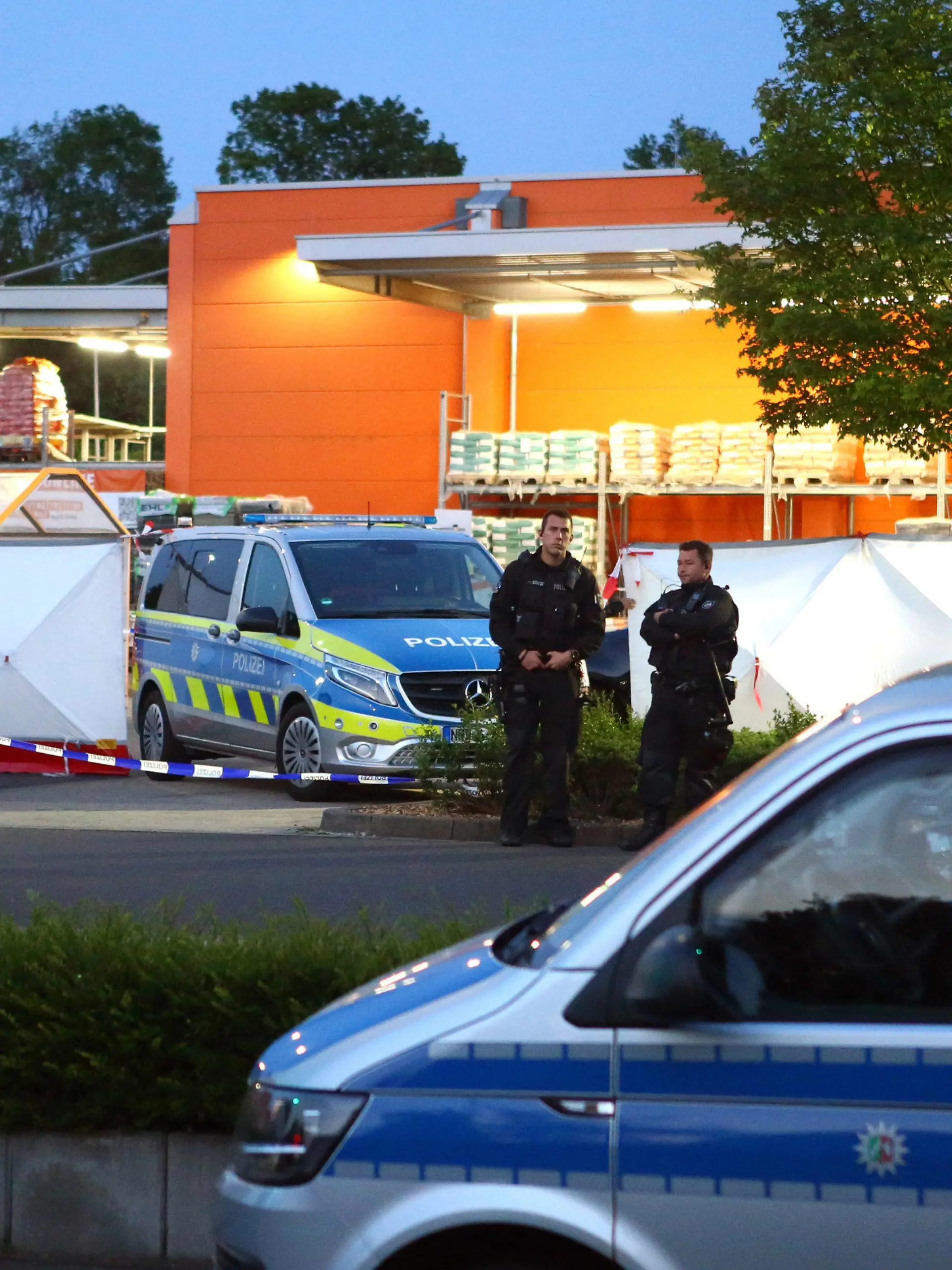 20.05.2023, Nordrhein-Westfalen, Aachen: Polizisten stehen am Tatort auf dem Parkplatz eines Baumarkts. In Aachen ist eine Frau am Samstagabend auf dem Parkplatz eines Baumarkts getötet worden. Die Polizei sperrte die Umgebung ab und setzte zur Fahndung einen Hubschrauber ein, wie ein Polizeisprecher am frühen Sonntagmorgen sagte. Schließlich wurde ein dringend Tatverdächtiger festgenommen. Die genauen Umstände und Hintergründe der Tat waren zunächst nicht bekannt. Eine Mordkommission übernahm die Ermittlungen zu dem Fall, über den zuvor die «Aachener Zeitung» berichtet hatte. Foto: Ralf Roeger/dpa - ACHTUNG: KfZ wurde aus rechtlichen Gründen gepixelt +++ dpa-Bildfunk +++