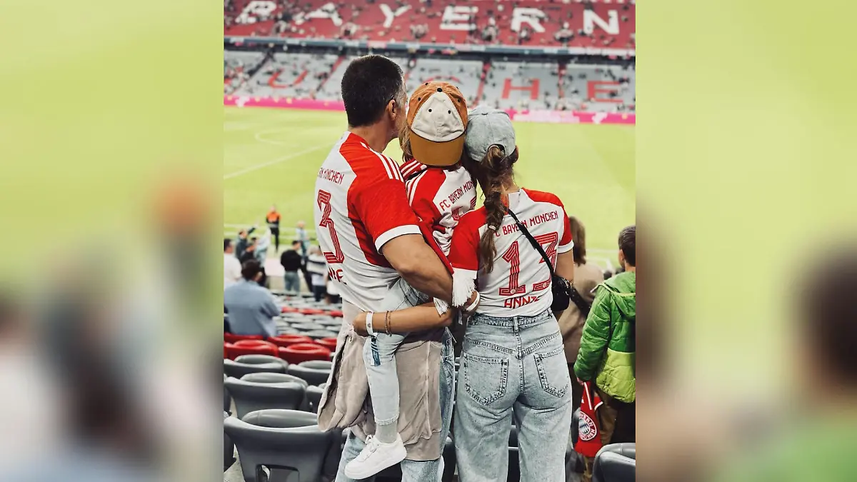 Wayne Carpendale steht neben seiner Frau, Annemarie Carpendale, im Fußballstadtion des FC Bayern München. Er trägt seinen Sohn Mads auf dem Arm. Man sieht die Familie von hinten, alle tragen Bayern-Trikots.