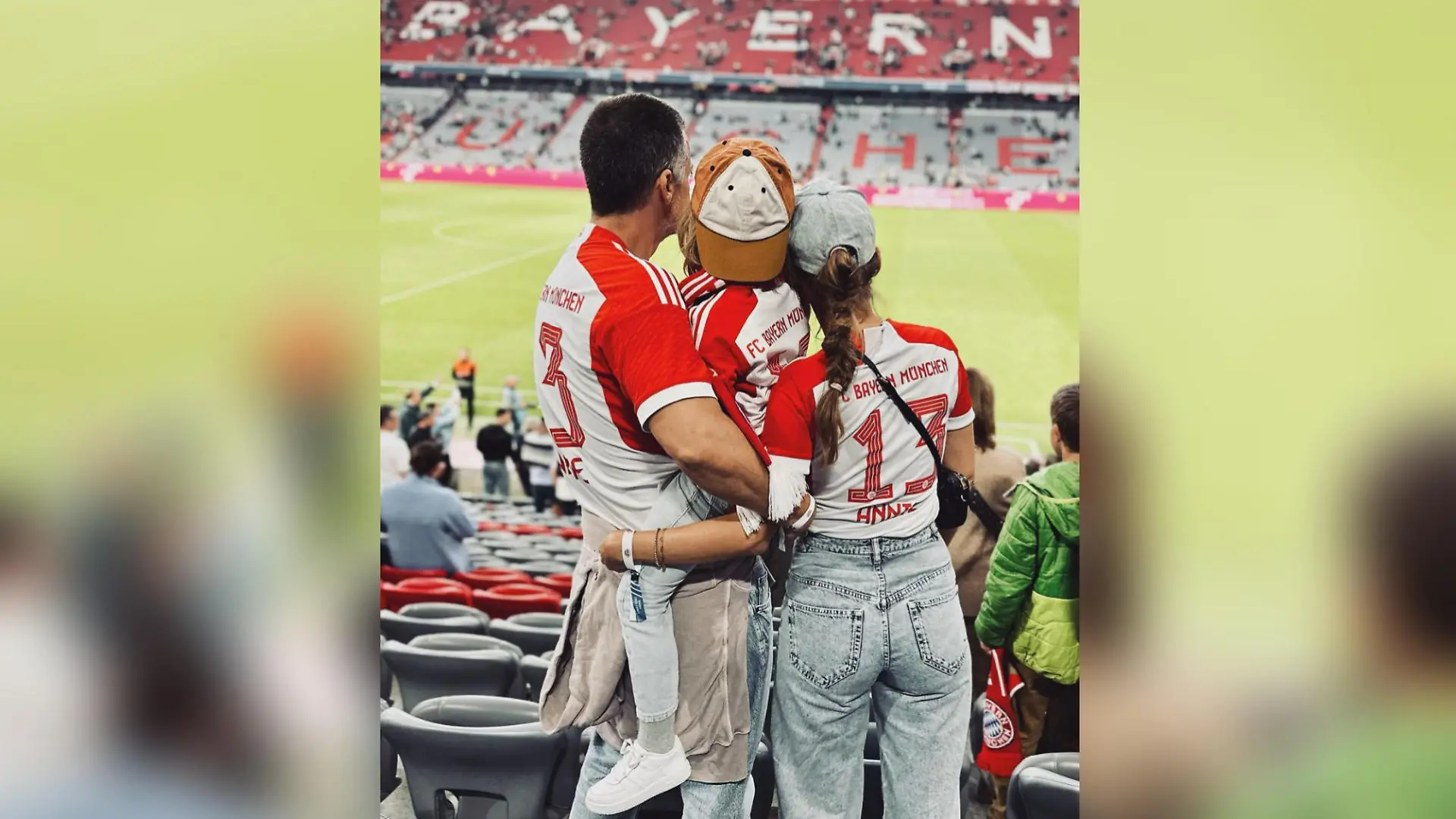 Wayne Carpendale steht neben seiner Frau, Annemarie Carpendale, im Fußballstadtion des FC Bayern München. Er trägt seinen Sohn Mads auf dem Arm. Man sieht die Familie von hinten, alle tragen Bayern-Trikots.