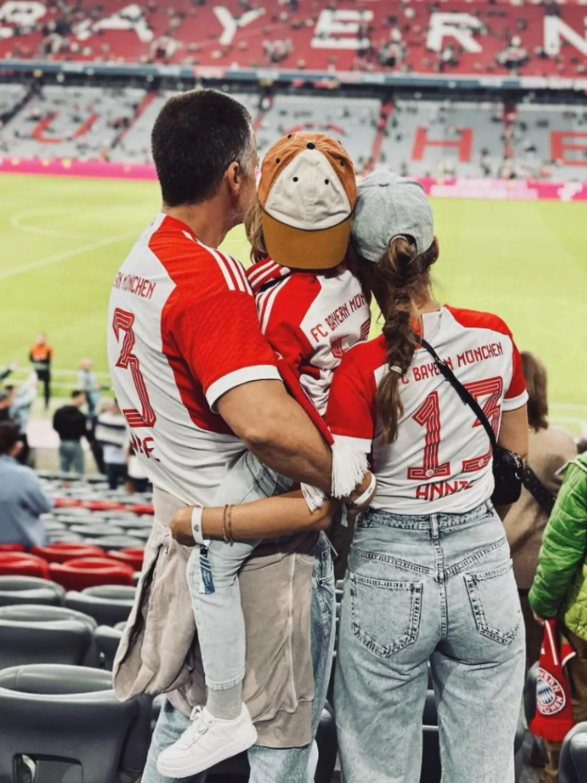 Wayne Carpendale steht neben seiner Frau, Annemarie Carpendale, im Fußballstadtion des FC Bayern München. Er trägt seinen Sohn Mads auf dem Arm. Man sieht die Familie von hinten, alle tragen Bayern-Trikots.