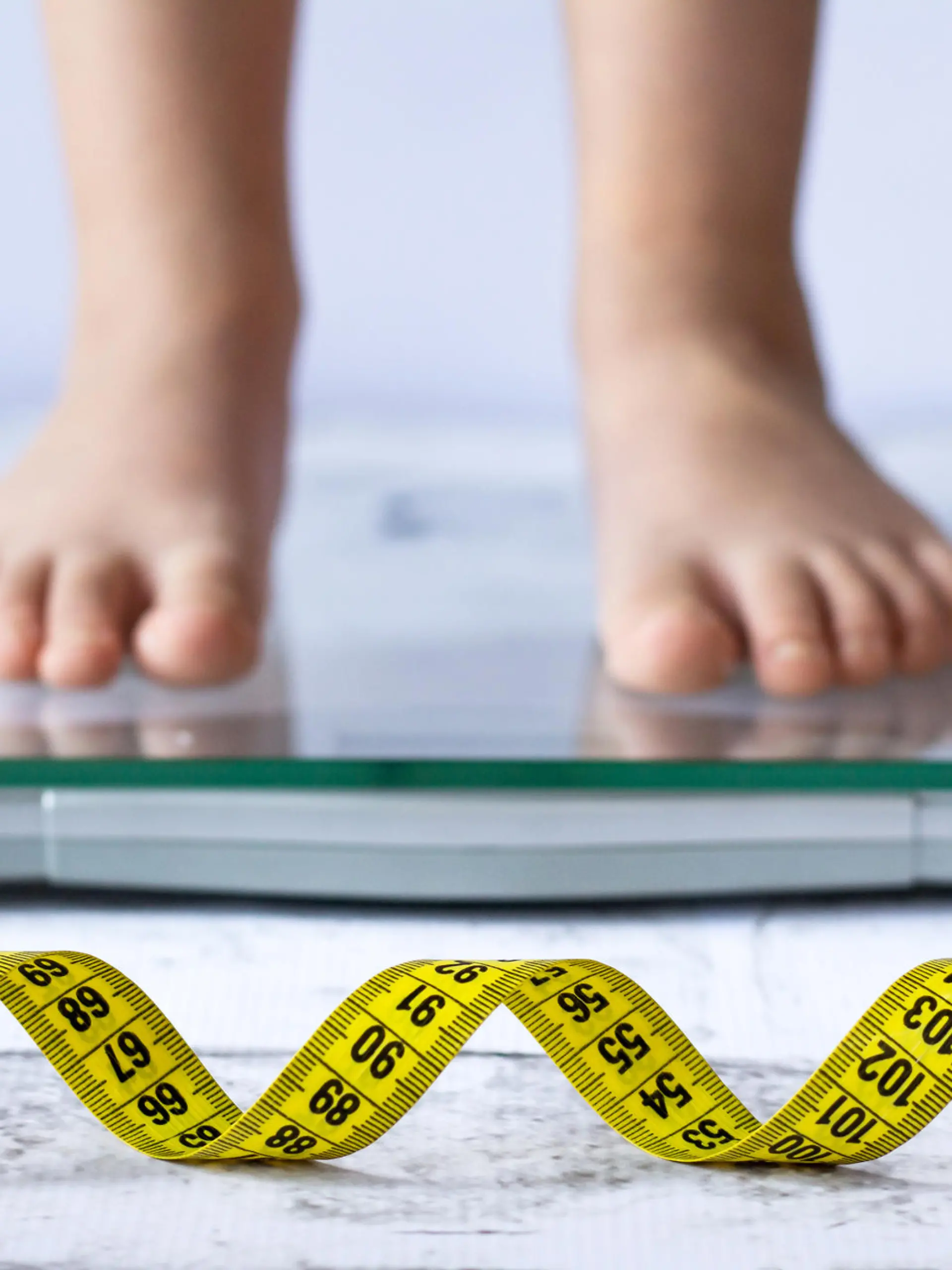 Weight control concept with centimeter in focus and blurred kid's feet on digital scale in the background. Child measuring weight, healthy growing.