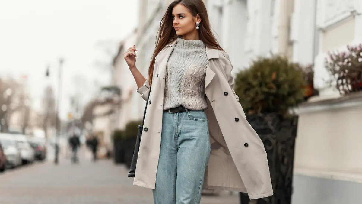 Model of a young beautiful woman in a fashion trench coat in a stylish knitted sweater with a leather black bag walks on the city near a white building. Elegant pretty trendy girl travels the street.