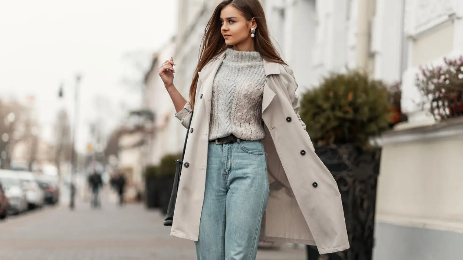 Model of a young beautiful woman in a fashion trench coat in a stylish knitted sweater with a leather black bag walks on the city near a white building. Elegant pretty trendy girl travels the street.