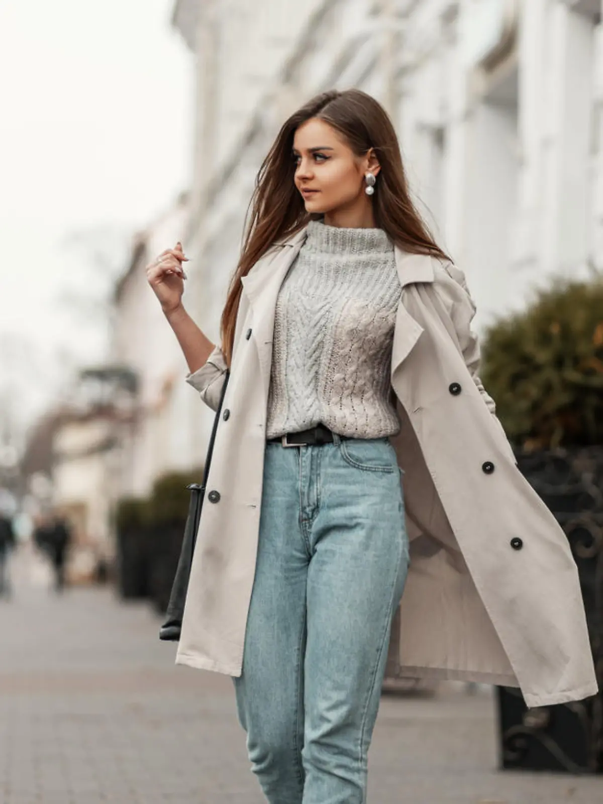 Model of a young beautiful woman in a fashion trench coat in a stylish knitted sweater with a leather black bag walks on the city near a white building. Elegant pretty trendy girl travels the street.