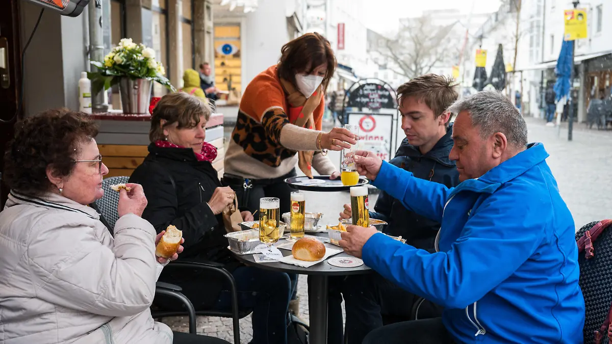 06.04.2021, Saarland, Saarbrücken: Wirtin Monika Müller (m) bringt ihren Gästen, die vor ihrer Kneipe «Glühwürmchen» in der Innenstadt sitzen, Getränke. Seit dem 06.04.2021 dürfen Gastronomen im Rahmen des Saarland Modells unter Auflagen ihre Außengastronomie wieder öffnen. Foto: Oliver Dietze/dpa +++ dpa-Bildfunk +++