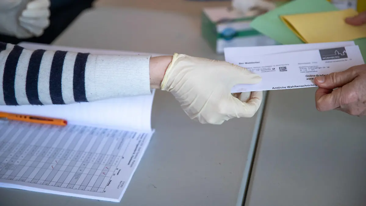 15.03.2020, Bayern, Nürnberg: Mit Schutzhandschuhen übernimmt zur Kommunalwahl in Bayern in einem Wahllokal ein Wahlhelfer eine amtliche Wahlbenachrichtigung. Insgesamt finden mehr als 4000 Wahlen in Bayern statt: Gemeinderäte, Stadträte, Kreistage. Zugleich gibt es in 24 der 25 kreisfreien Städte die Wahl des Oberbürgermeisters und in 64 der 71 Landkreise die Wahl des Landrats. Bayernweit sind fast 40 000 Mandate zu vergeben. Für die Parteien sind die Kommunalwahlen ein weiterer Stimmungstest nach der Landtagswahl 2018 und der Europawahl 2019. Foto: Daniel Karmann/dpa +++ dpa-Bildfunk +++