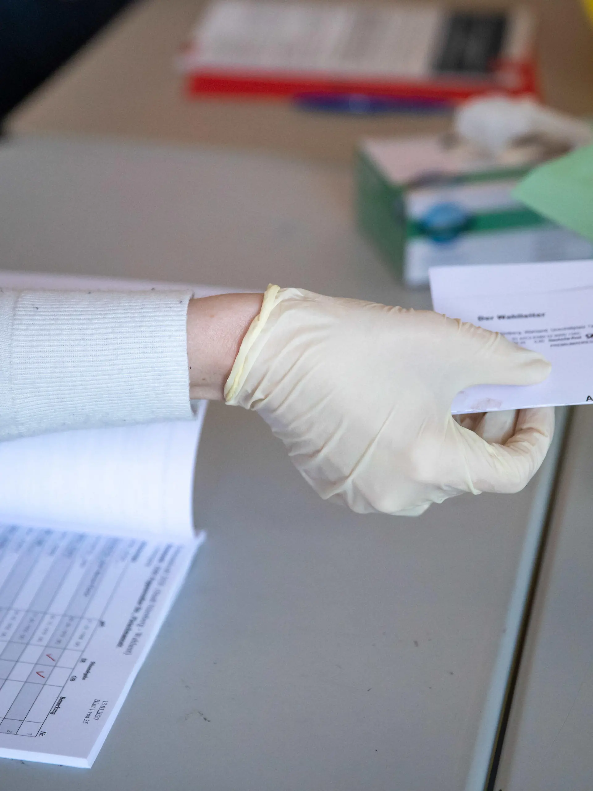 15.03.2020, Bayern, Nürnberg: Mit Schutzhandschuhen übernimmt zur Kommunalwahl in Bayern in einem Wahllokal ein Wahlhelfer eine amtliche Wahlbenachrichtigung. Insgesamt finden mehr als 4000 Wahlen in Bayern statt: Gemeinderäte, Stadträte, Kreistage. Zugleich gibt es in 24 der 25 kreisfreien Städte die Wahl des Oberbürgermeisters und in 64 der 71 Landkreise die Wahl des Landrats. Bayernweit sind fast 40 000 Mandate zu vergeben. Für die Parteien sind die Kommunalwahlen ein weiterer Stimmungstest nach der Landtagswahl 2018 und der Europawahl 2019. Foto: Daniel Karmann/dpa +++ dpa-Bildfunk +++