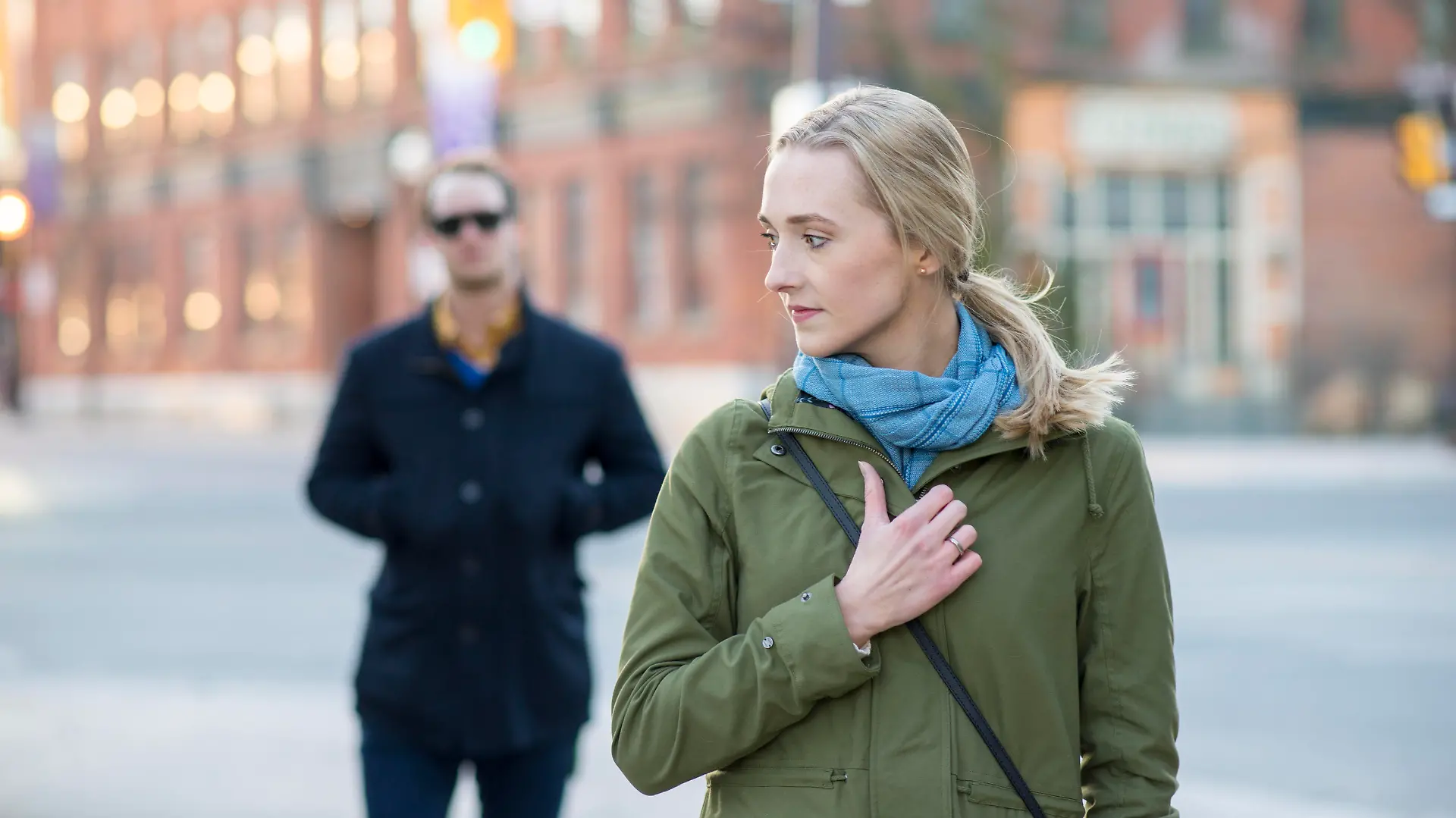 Scared young woman with blonde hair looking back over her shoulder at a stranger in a black trench coat that is following behind her downtown in an urban city.