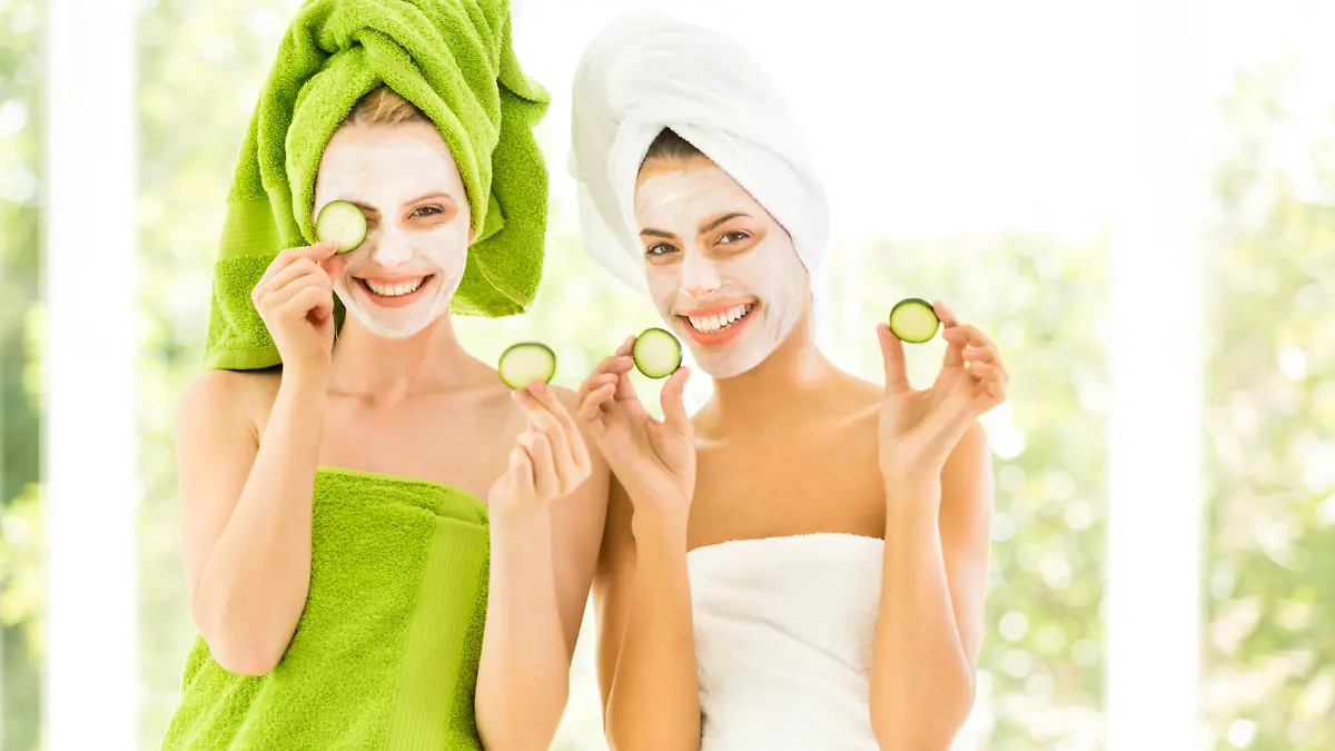 Two smiling young women with towel on head and slice of cucumber in hands.