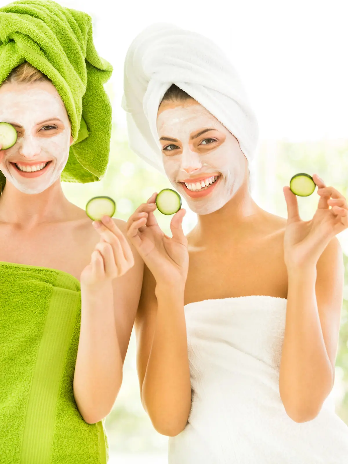 Two smiling young women with towel on head and slice of cucumber in hands.
