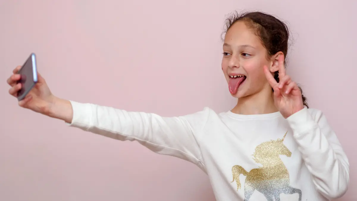 Young brunette girl, making selfie, showing v-sign and tongue out, funny face, posing at pink background. Soft focus on face of child.