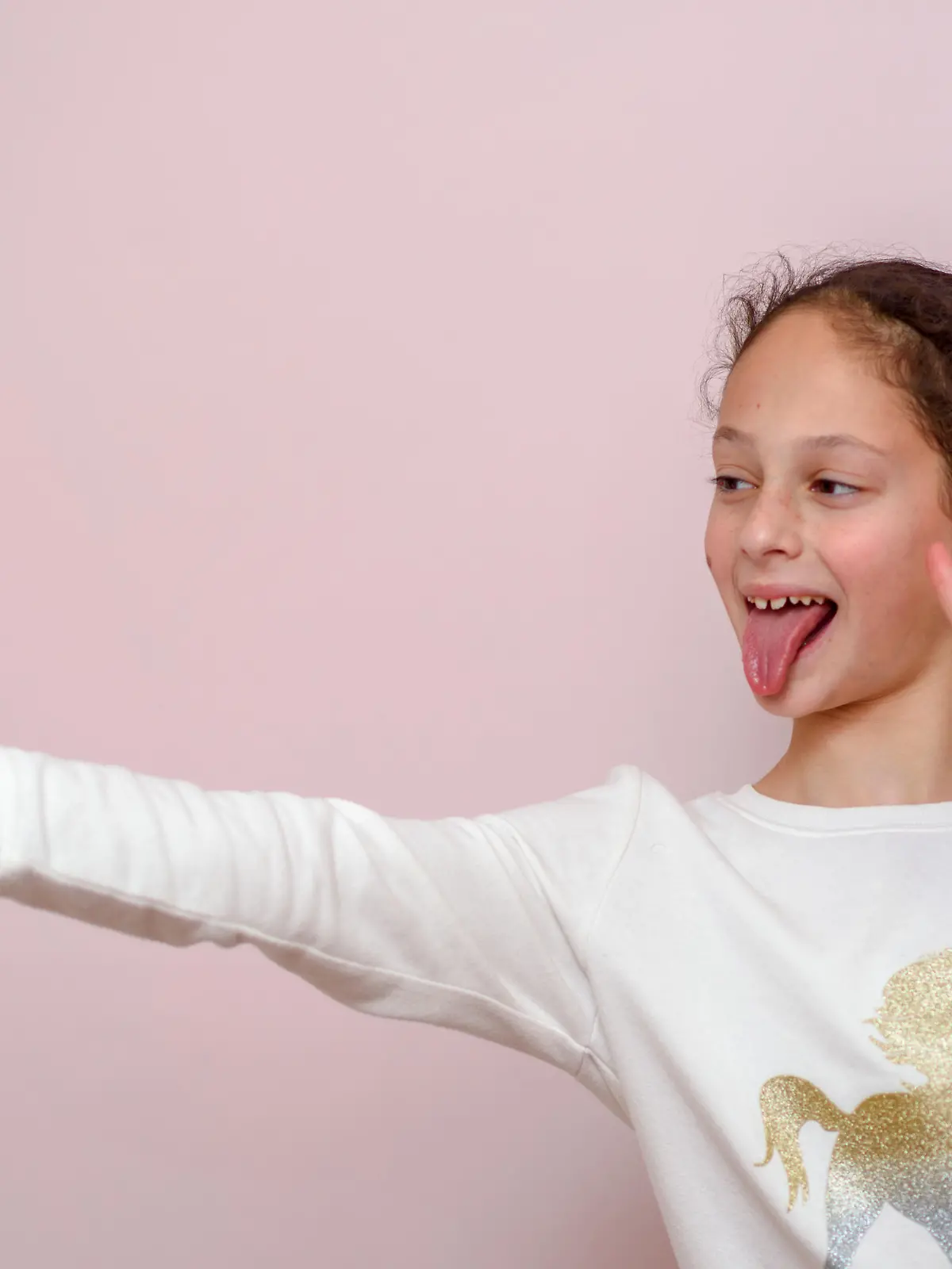 Young brunette girl, making selfie, showing v-sign and tongue out, funny face, posing at pink background. Soft focus on face of child.