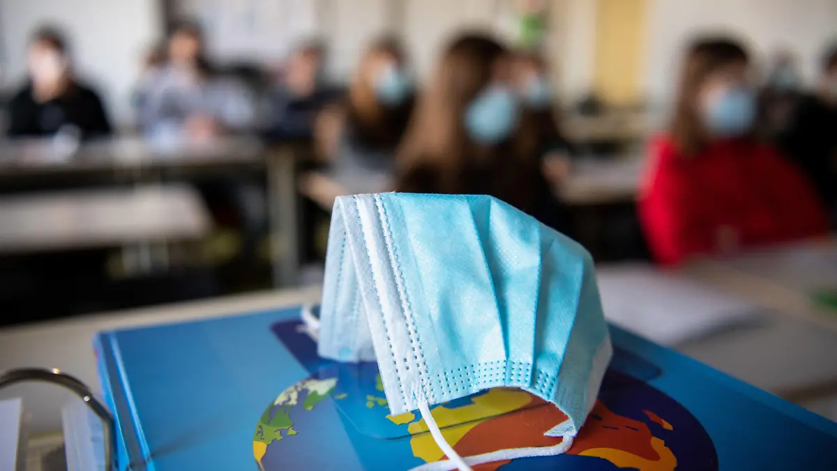 ARCHIV - 17.11.2020, Bayern, München: ILLUSTRATION - Ein Mund- und Nasentschutz liegt im Unterricht in einem Geographie-Seminar in der Jahrgangsstufe elf am staatlichen Gymnasium Trudering auf einem Weltalas, während im Hintergrund die Schülerinnen und Schüler mit Mund- und Nasenschutz zu sehen sind. (zu dpa «Mathe und Deutsch mit Maske: Schulen öffnen für den Corona-Alltag») Foto: Matthias Balk/dpa +++ dpa-Bildfunk +++