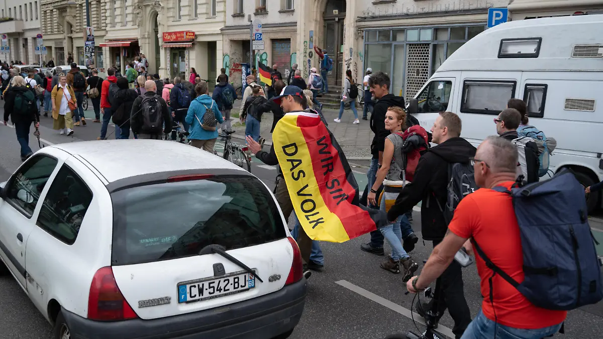 Aus Protest gegen die Corona-Politik wandern Demonstranten durch eine Straße. Ein Mann trägt eine Deutschland-Fahne mit der Aufschrift "Wir sind das Volk" auf dem Rücken. Neun Demonstrationen waren von der Polizei verboten worden, darunter Kundgebungen der «Initiative Querdenken» auf der Straße des 17. Juni. Eine für Samstag und Sonntag angemeldete Versammlung mit je 500 erwarteten Teilnehmern darf nun doch stattfinden.