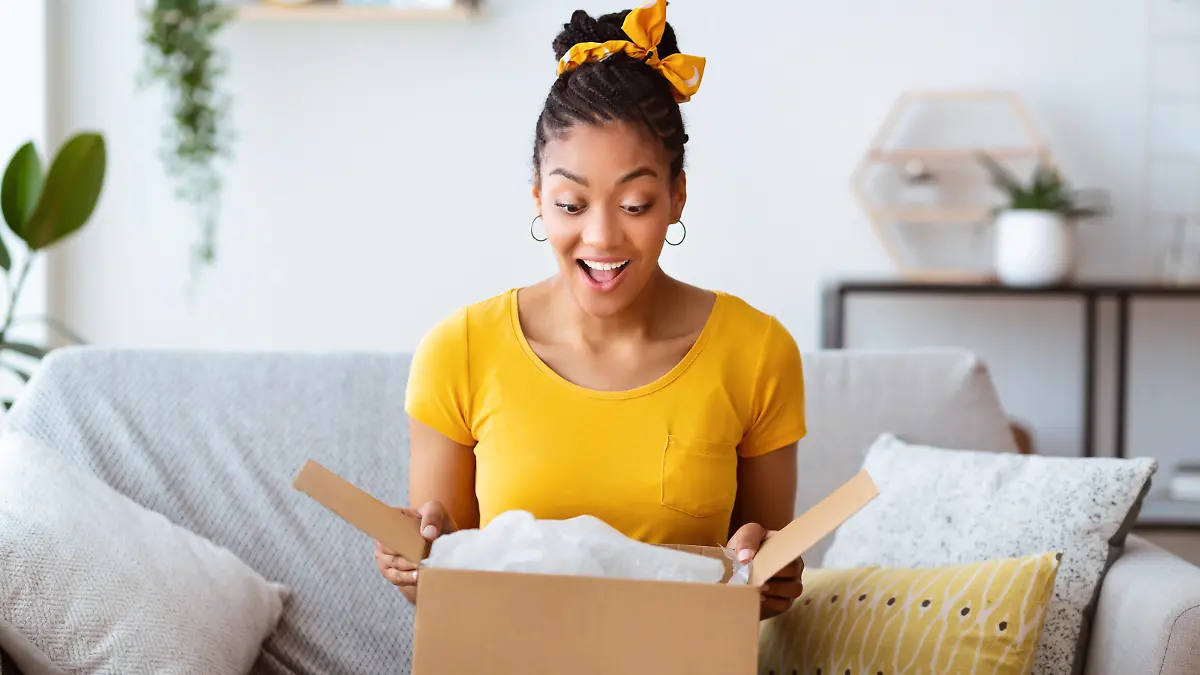 Buying Via Internet. Excited afro girl sitting on sofa unboxing cardboard delivery package, copy space