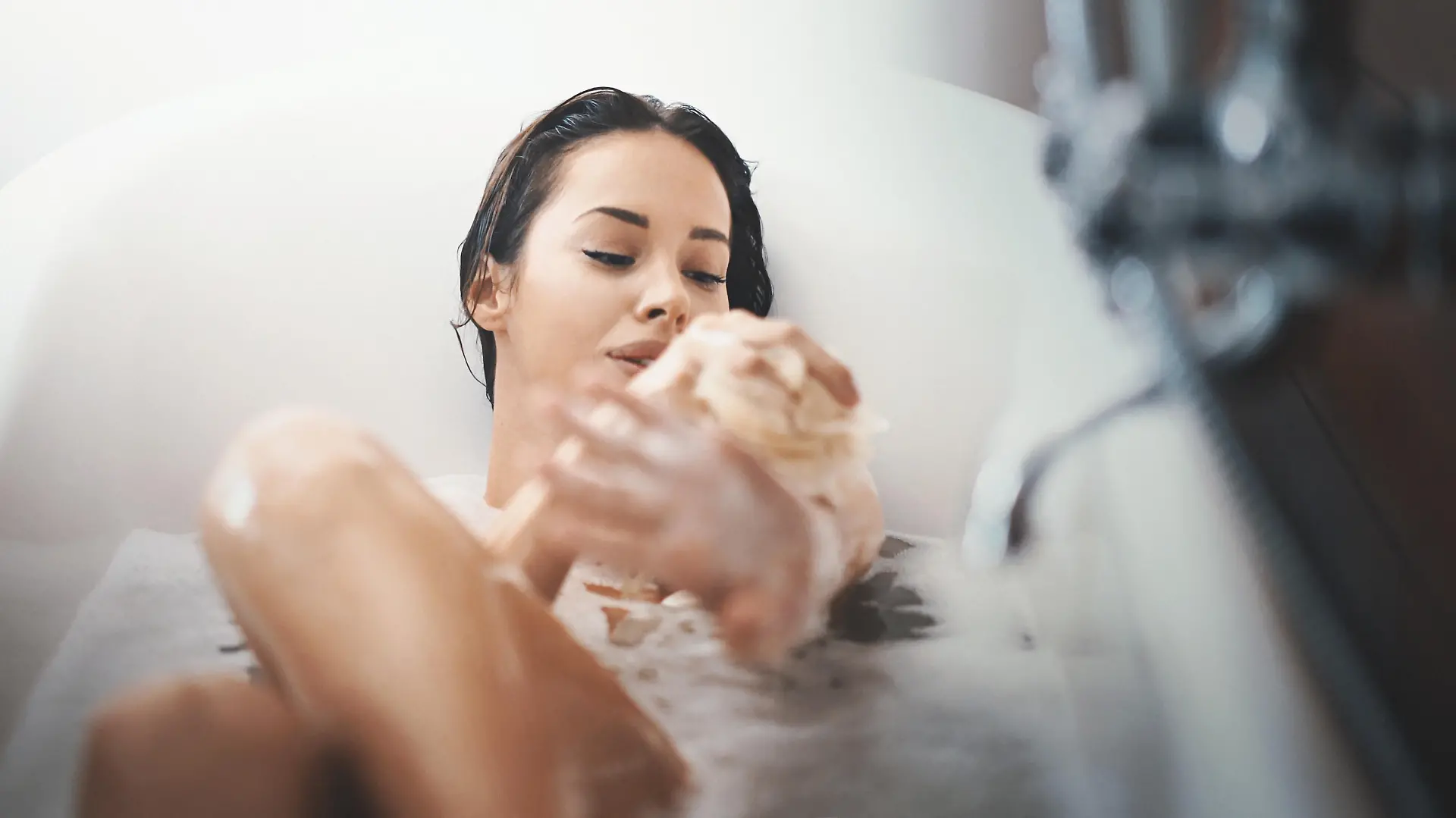 Closeup of attractive mid 20's woman taking a long bath in a bathtub. She's slowly scrubbing off with a bath sponge and not planning to get out some time soon.