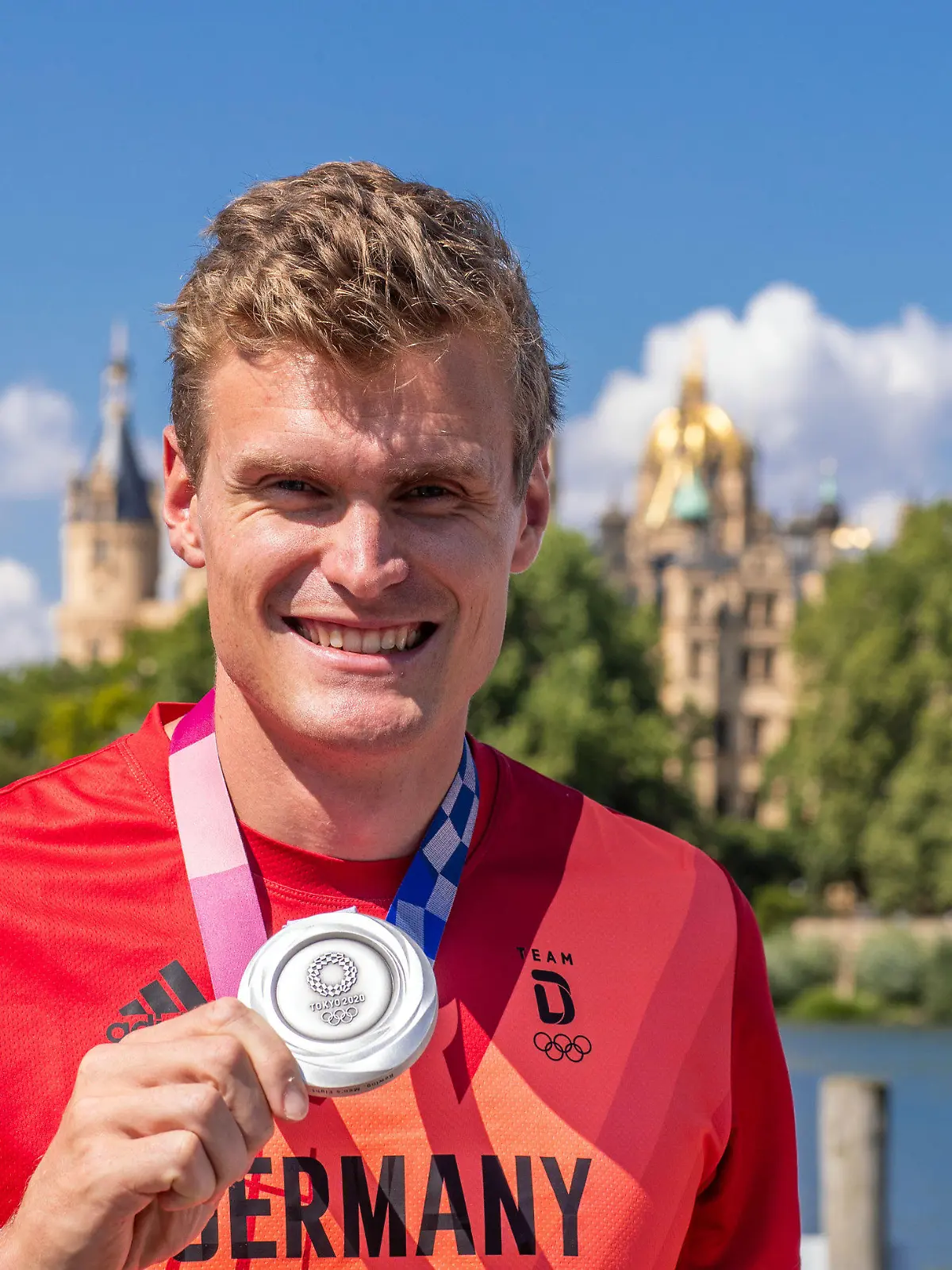 06.08.2021, Mecklenburg-Vorpommern, Schwerin: Hannes Ocik, Schlagmann im deutschen Ruderachter bei den Olympischen Spielen in Tokio, steht mit seiner Silbermedaille beim Empfang vor dem Schweriner Schloß. Ocik wurde im Ruderverein Schwerin nach seiner Rückkehr aus Japan begrüßt. Foto: Jens Büttner/dpa-Zentralbild/dpa +++ dpa-Bildfunk +++