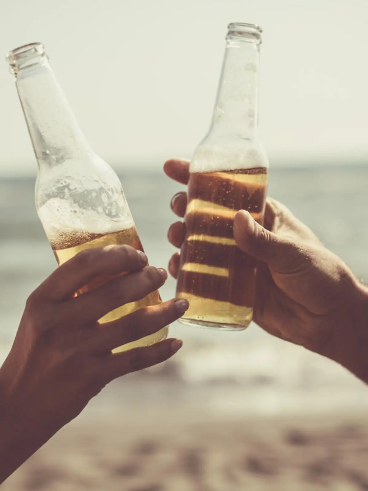 Close-up shot of male and female hand doing celebratory toast with beer bottles at the beach.