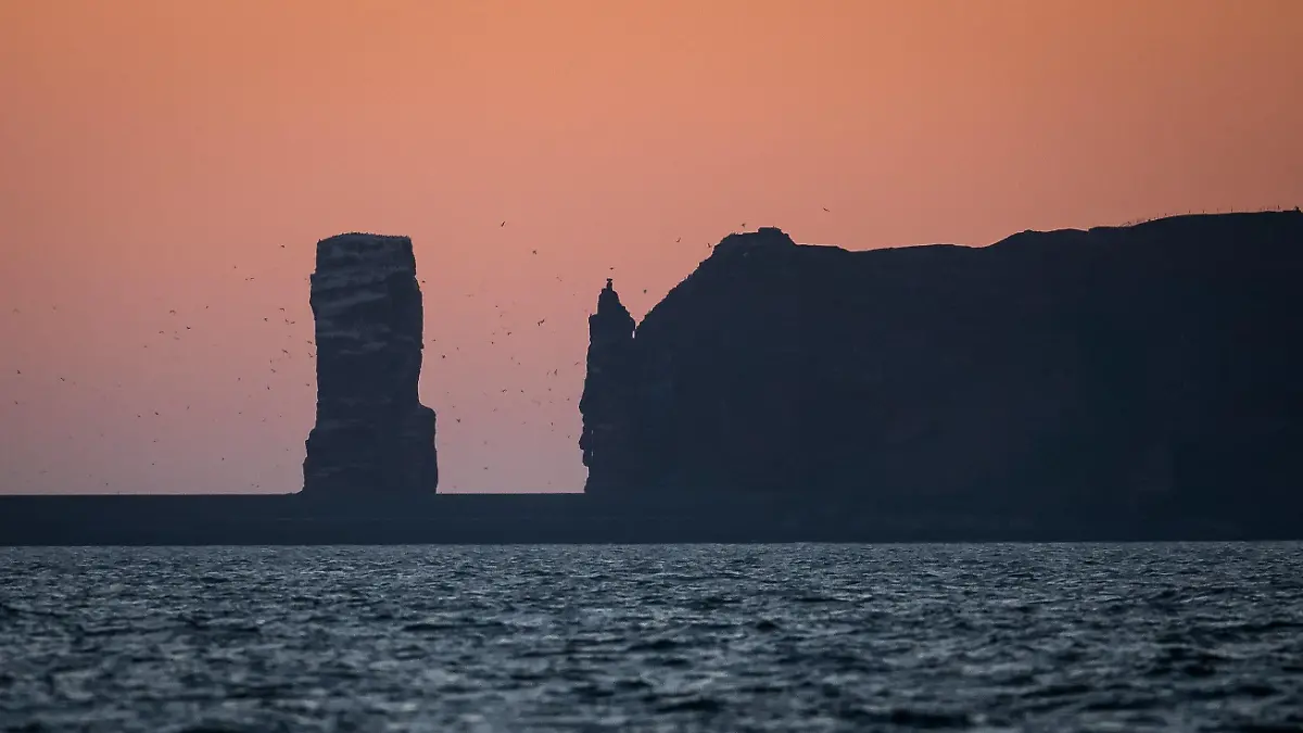 23.04.2020, Schleswig-Holstein, Helgoland: Die Sonne geht hinter den Klippen von Helgoland auf. Startschuss für ein großes maritimes Erprobungsfeld vor Helgoland: Wissenschaftler des Testzentrums für maritime Technologien haben mit Tonnen ein drei Millionen Quadratmeter großes Areal unweit der Nordseeinsel abgesteckt. Es soll künftig Erprobungsmöglichkeiten für maritime Robotik bieten. Hersteller und Wissenschaftler können dort unterschiedliche Szenarien erproben. Beispielsweise können autonome Unterwasserfahrzeuge für Offshore-Windkraftanlagen unter realen Bedingungen im Meer oder über das Wasser fliegende Drohnen für Inspektionsarbeiten erprobt werden. Foto: Sina Schuldt/dpa +++ dpa-Bildfunk +++