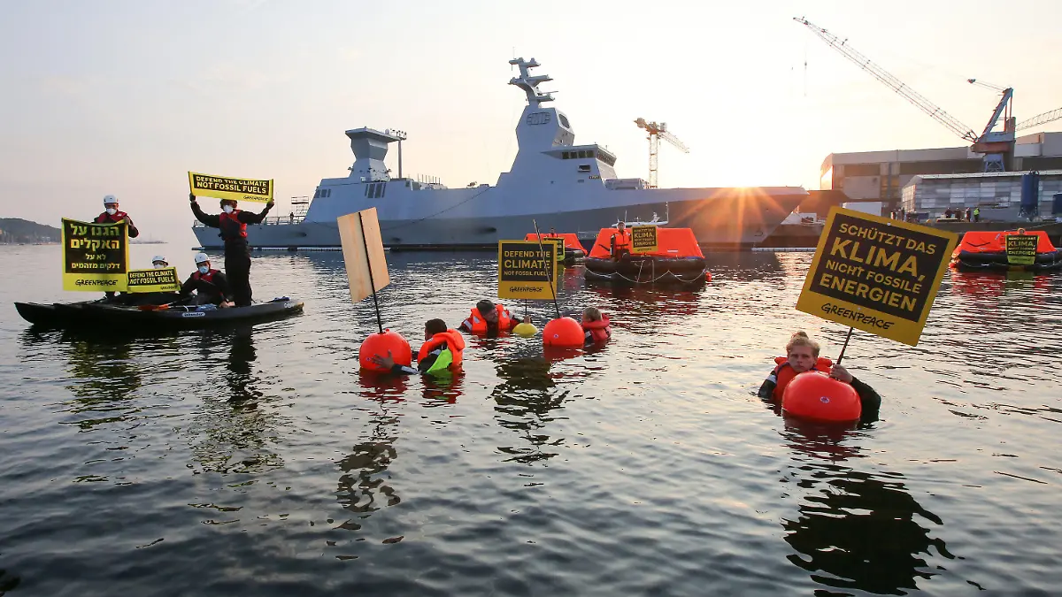 12.08.2021, Schleswig-Holstein, Kiel: Greenpeace-Aktivisten halten bei einer Aktion gegen Ausbeutung fossiler Energien Schilder mit Aufschriften in verschiedenen Sprachen. Im Hintergrund ist ein israelisches Kriegsschiff, die Korvette I.N.S. Nitzachon, zu sehen. Greenpeace-Aktivisten aus Israel und Deutschland haben am Donnerstagmorgen gemeinsam im Kieler Hafen gegen die Ausbeutung fossiler Energien demonstriert. Eigenen Angaben zufolge protestierten 13 Menschen gegen das Auslaufen eines Schiffes und zogen mit fünf Rettungsinseln, vier Kajaks und vier Schwimmenden eine Linie vor der Korvette. Foto: Bodo Marks/dpa +++ dpa-Bildfunk +++