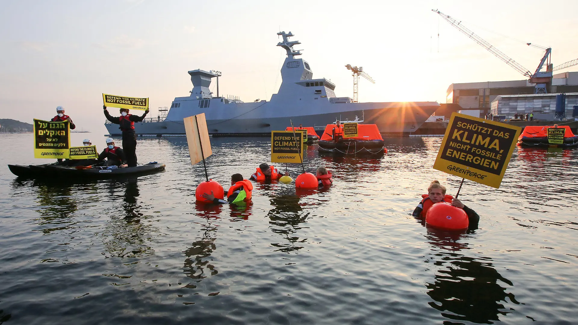 12.08.2021, Schleswig-Holstein, Kiel: Greenpeace-Aktivisten halten bei einer Aktion gegen Ausbeutung fossiler Energien Schilder mit Aufschriften in verschiedenen Sprachen. Im Hintergrund ist ein israelisches Kriegsschiff, die Korvette I.N.S. Nitzachon, zu sehen. Greenpeace-Aktivisten aus Israel und Deutschland haben am Donnerstagmorgen gemeinsam im Kieler Hafen gegen die Ausbeutung fossiler Energien demonstriert. Eigenen Angaben zufolge protestierten 13 Menschen gegen das Auslaufen eines Schiffes und zogen mit fünf Rettungsinseln, vier Kajaks und vier Schwimmenden eine Linie vor der Korvette. Foto: Bodo Marks/dpa +++ dpa-Bildfunk +++