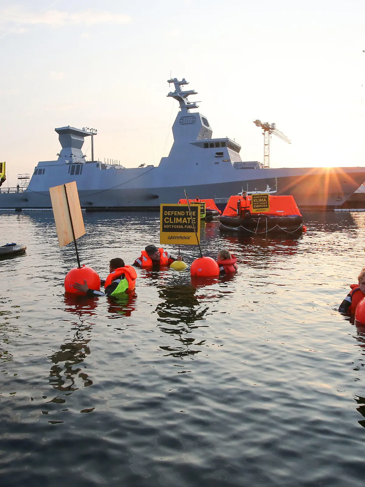 12.08.2021, Schleswig-Holstein, Kiel: Greenpeace-Aktivisten halten bei einer Aktion gegen Ausbeutung fossiler Energien Schilder mit Aufschriften in verschiedenen Sprachen. Im Hintergrund ist ein israelisches Kriegsschiff, die Korvette I.N.S. Nitzachon, zu sehen. Greenpeace-Aktivisten aus Israel und Deutschland haben am Donnerstagmorgen gemeinsam im Kieler Hafen gegen die Ausbeutung fossiler Energien demonstriert. Eigenen Angaben zufolge protestierten 13 Menschen gegen das Auslaufen eines Schiffes und zogen mit fünf Rettungsinseln, vier Kajaks und vier Schwimmenden eine Linie vor der Korvette. Foto: Bodo Marks/dpa +++ dpa-Bildfunk +++