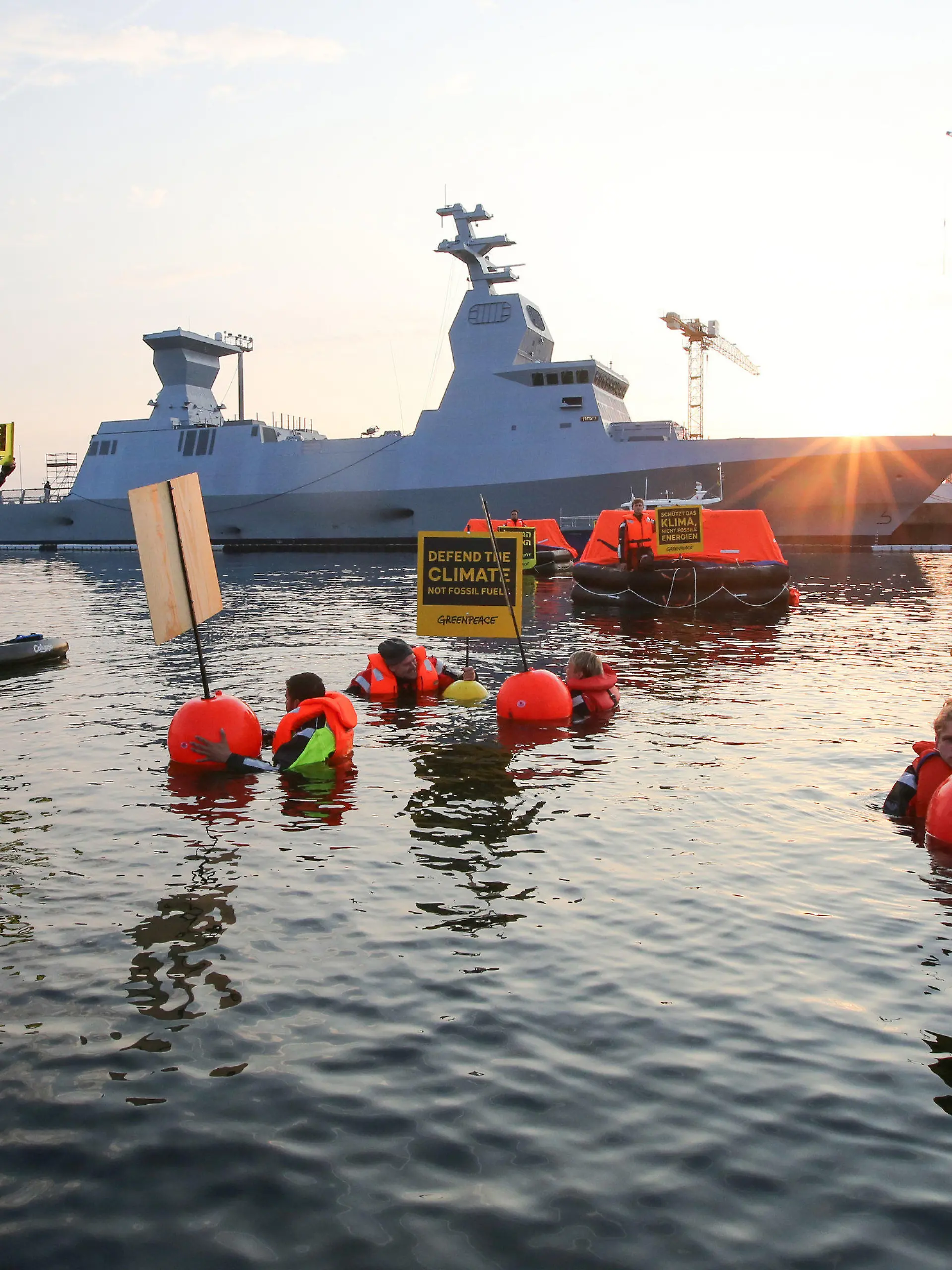 12.08.2021, Schleswig-Holstein, Kiel: Greenpeace-Aktivisten halten bei einer Aktion gegen Ausbeutung fossiler Energien Schilder mit Aufschriften in verschiedenen Sprachen. Im Hintergrund ist ein israelisches Kriegsschiff, die Korvette I.N.S. Nitzachon, zu sehen. Greenpeace-Aktivisten aus Israel und Deutschland haben am Donnerstagmorgen gemeinsam im Kieler Hafen gegen die Ausbeutung fossiler Energien demonstriert. Eigenen Angaben zufolge protestierten 13 Menschen gegen das Auslaufen eines Schiffes und zogen mit fünf Rettungsinseln, vier Kajaks und vier Schwimmenden eine Linie vor der Korvette. Foto: Bodo Marks/dpa +++ dpa-Bildfunk +++