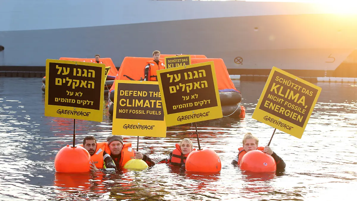 12.08.2021, Schleswig-Holstein, Kiel: Greepeace-Aktivisten halten bei einer Aktion gegen Ausbeutung fossiler Energien Schilder mit Aufschriften in verschiedenen Sprachen. Im Hintergrund ist ein Teil eines israelisches Kriegsschiffs, die Korvette I.N.S. Nitzachon, zu sehen. Greenpeace-Aktivisten aus Israel und Deutschland haben am Donnerstagmorgen gemeinsam im Kieler Hafen gegen die Ausbeutung fossiler Energien demonstriert. Eigenen Angaben zufolge protestierten 13 Menschen gegen das Auslaufen eines Schiffes und zogen mit fünf Rettungsinseln, vier Kajaks und vier Schwimmenden eine Linie vor der Korvette. Foto: Bodo Marks/dpa +++ dpa-Bildfunk +++