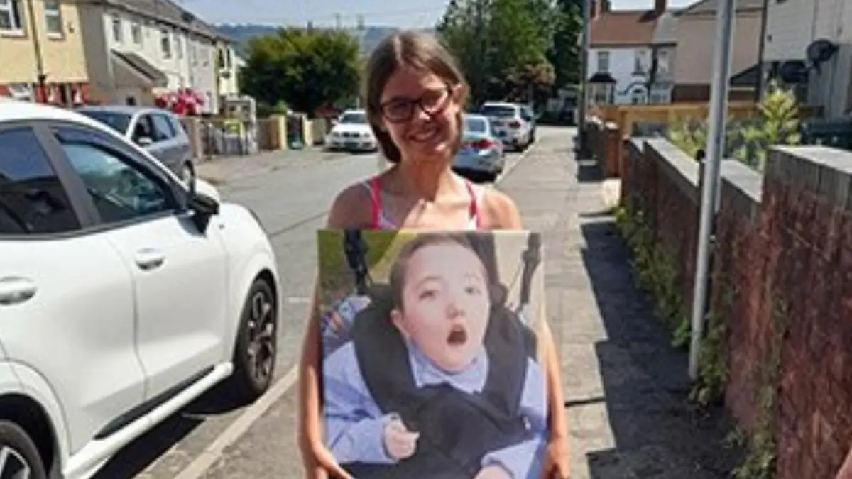 Mädchen mit Brille steht auf einem Fußweg und hat ein Schild um dem Bauch, auf dem ihr Bruder zu sehen ist.