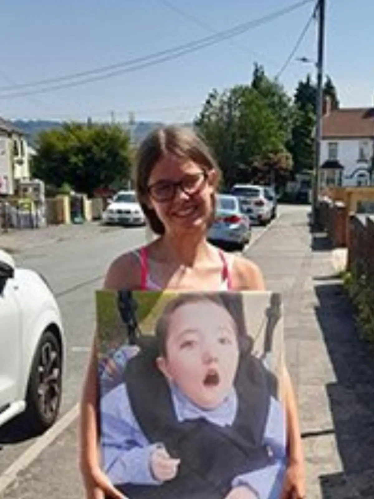 Mädchen mit Brille steht auf einem Fußweg und hat ein Schild um dem Bauch, auf dem ihr Bruder zu sehen ist.