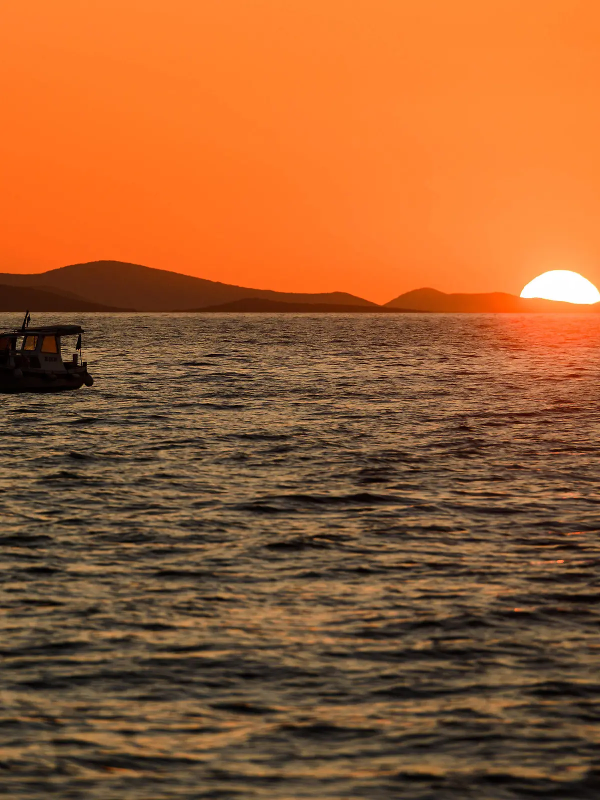 Sonnenuntergang am Meer, Sonne verschwindet hinter einem Berg am Horizont, im Vordergrund sieht man die Umrisse eines Schiffes auf dem Meer.