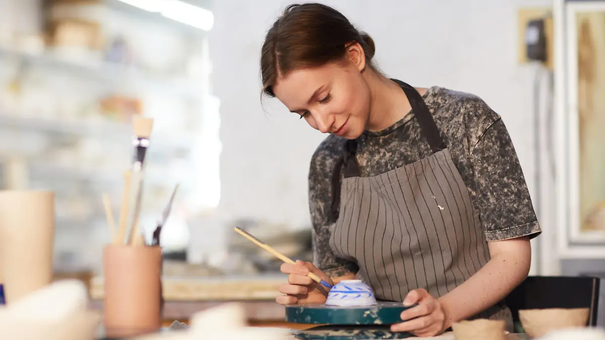 Content attractive skilled young lady in apron standing at table and drawing blue leaves on ceramic bowl in pottery workshop