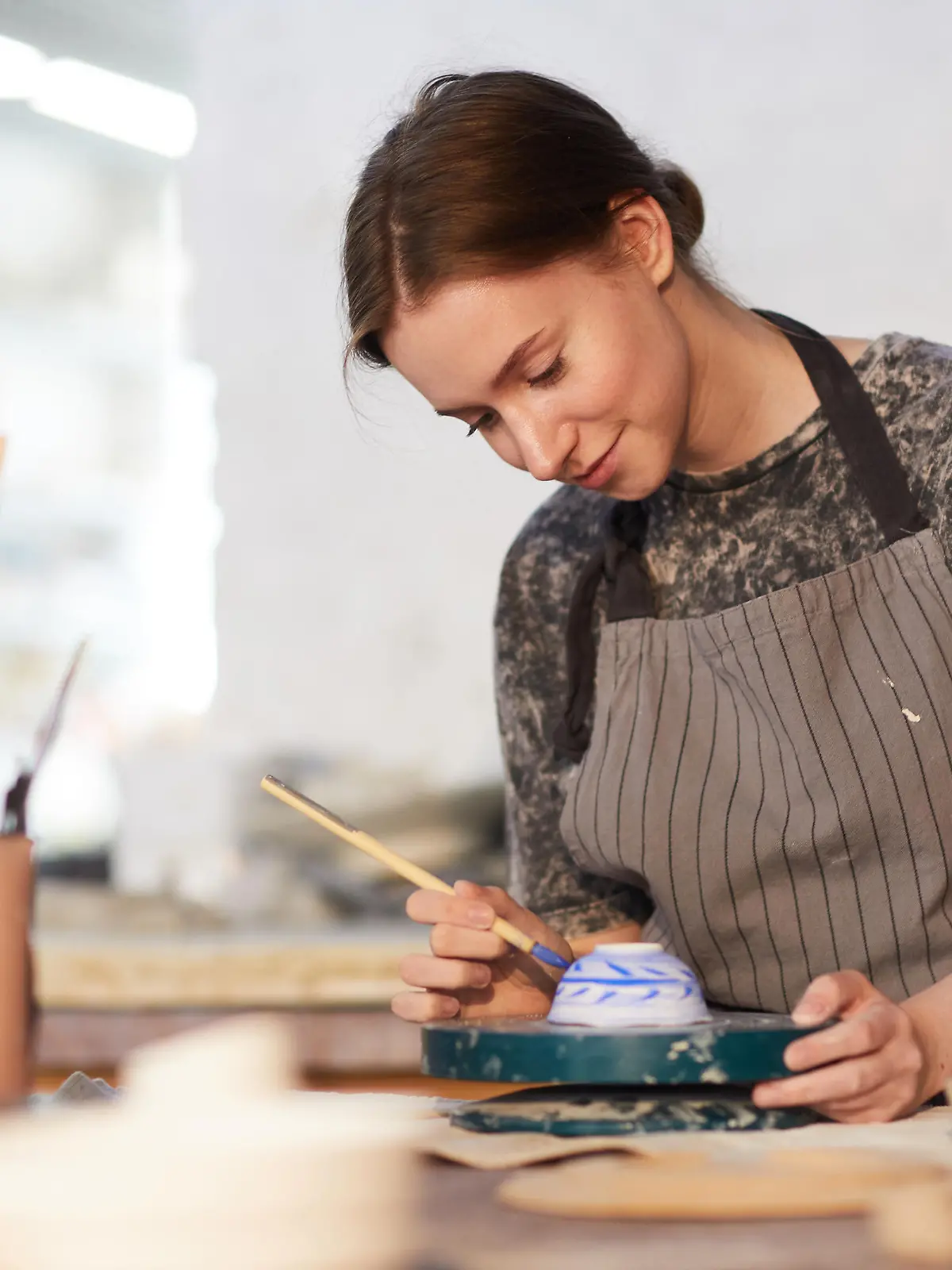 Content attractive skilled young lady in apron standing at table and drawing blue leaves on ceramic bowl in pottery workshop