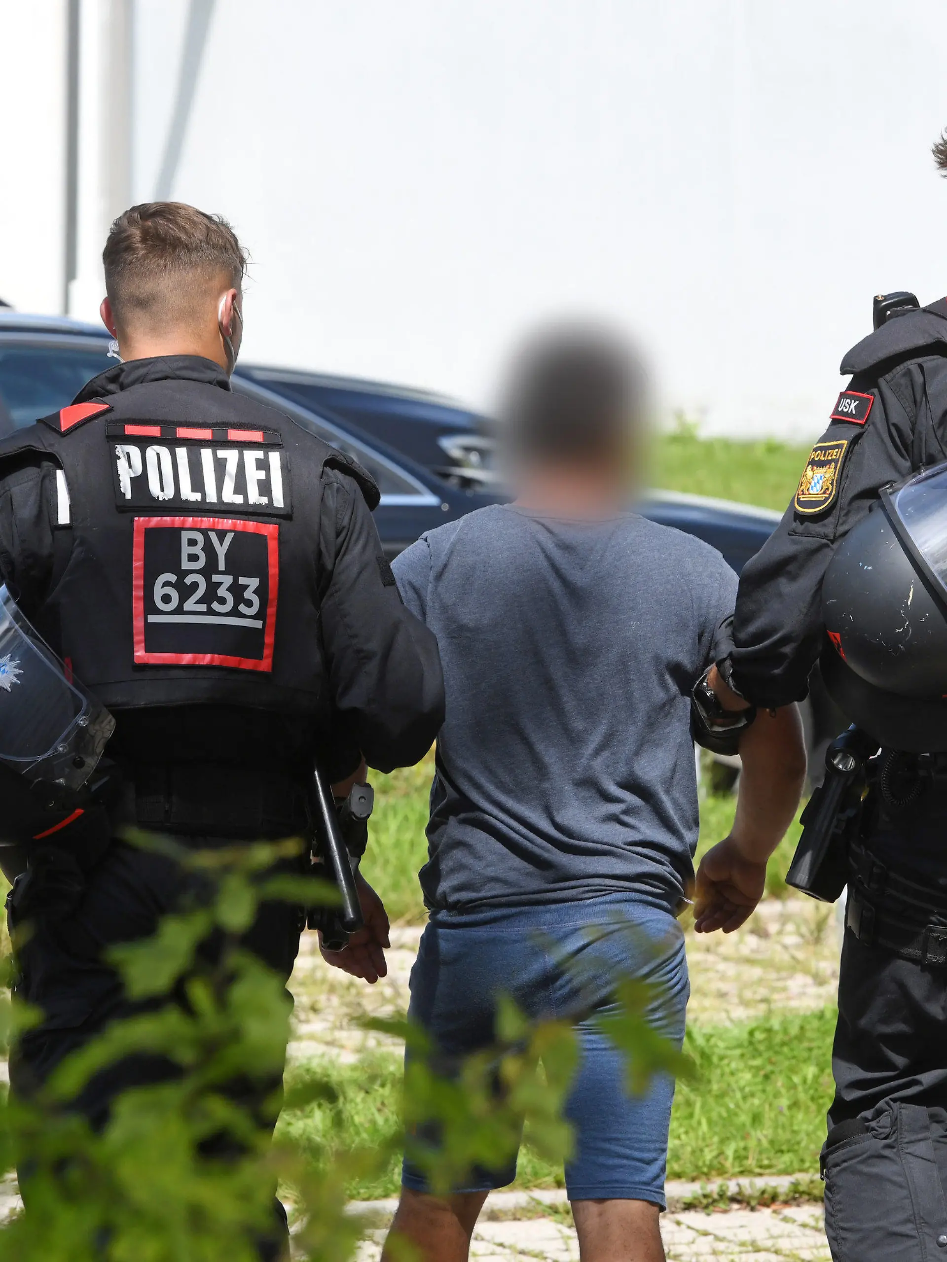 10.08.2021, Bayern, Miesbach: Zwei Einsatzkräfte der Polizei führen vor der Polizeiinspektion einen Tatverdächtigen ab. Mitglieder einer Großfamilie wollten sich Zugang zur Polizeistation verschaffen und griffen die Beamten mit Steinen an. Vorausgegangen war die Festnahme einer Person wegen Verdachts auf ein Sexualdelikt. Foto: Tobias Hase/dpa - ACHTUNG: Der Tatverdächtige wurde aus rechtlichen Gründen gepixelt +++ dpa-Bildfunk +++