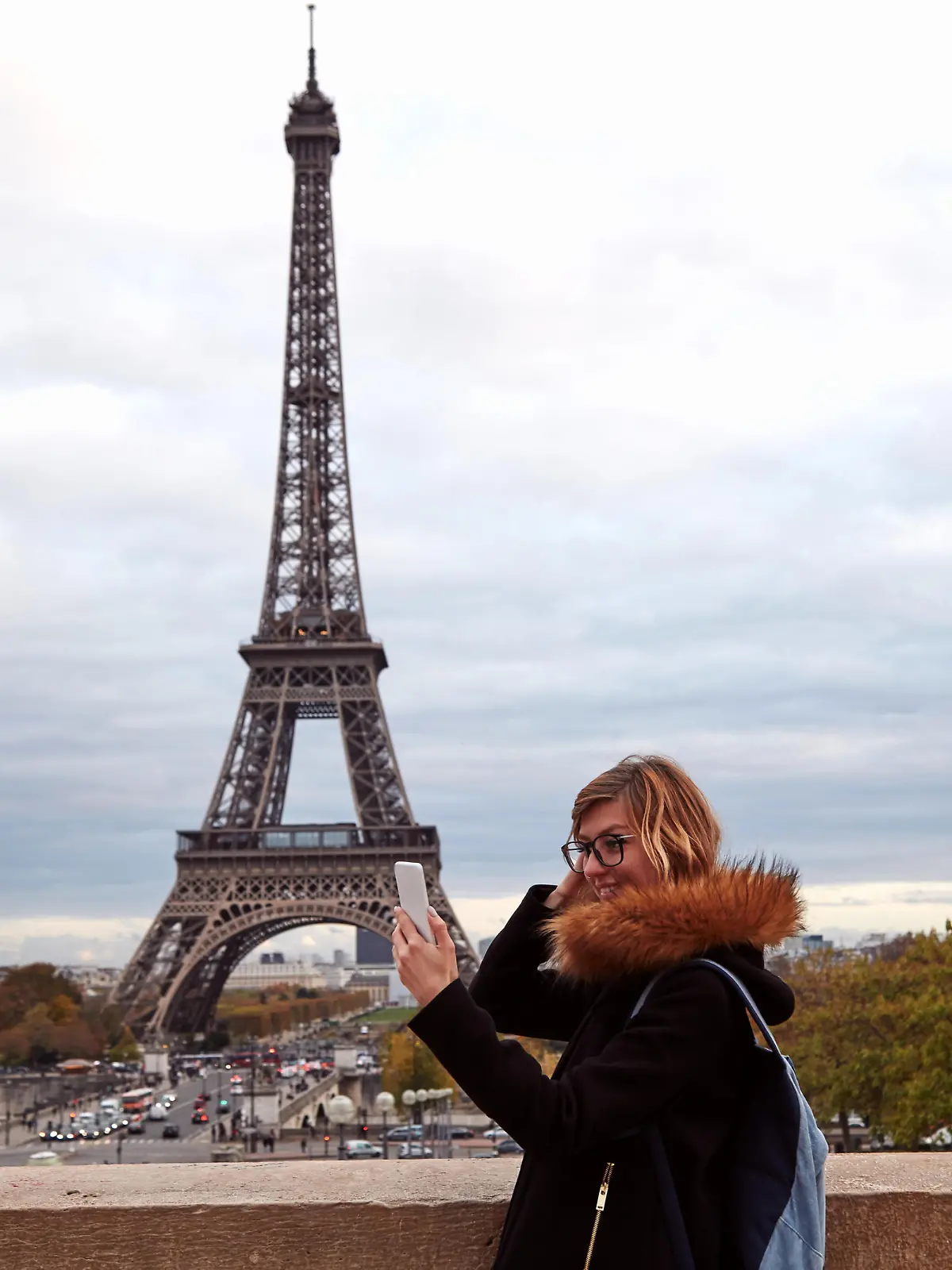 Eine junge Frau steht vor dem Eiffelturm und schaut auf ihr Handy.