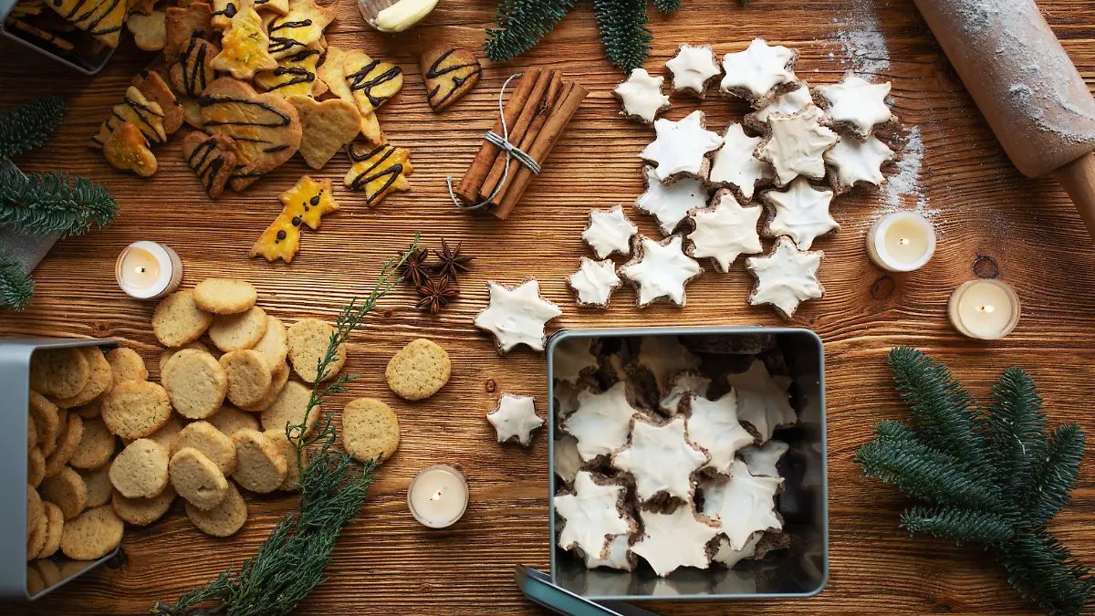 Verschiedene Weihnachtskekse, die verteilt auf einem Holztisch liegen, vereinzelt auch in Keksdosen