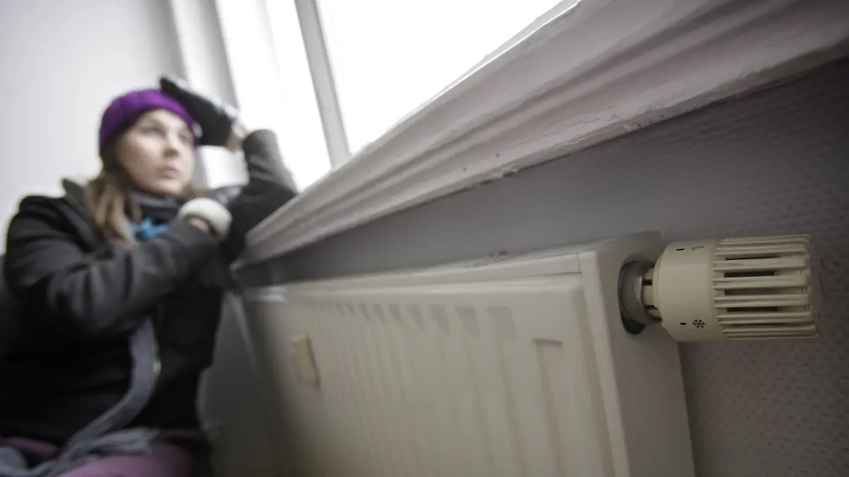 Junge Frau sitzt frierend an der Heizung mit Mütze und Handschuhe vor einem offenen Fenster, dessen Rollladen nicht geschlossen isnd