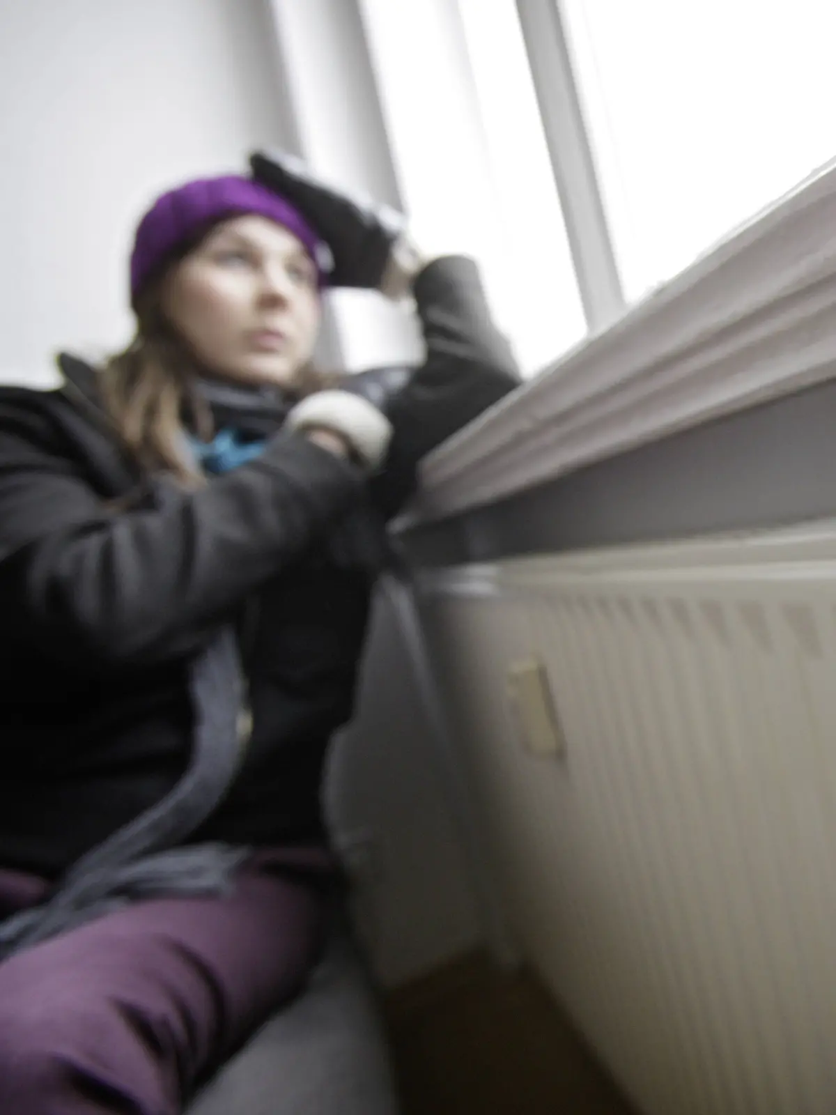 Junge Frau sitzt frierend an der Heizung mit Mütze und Handschuhe vor einem offenen Fenster, dessen Rollladen nicht geschlossen isnd