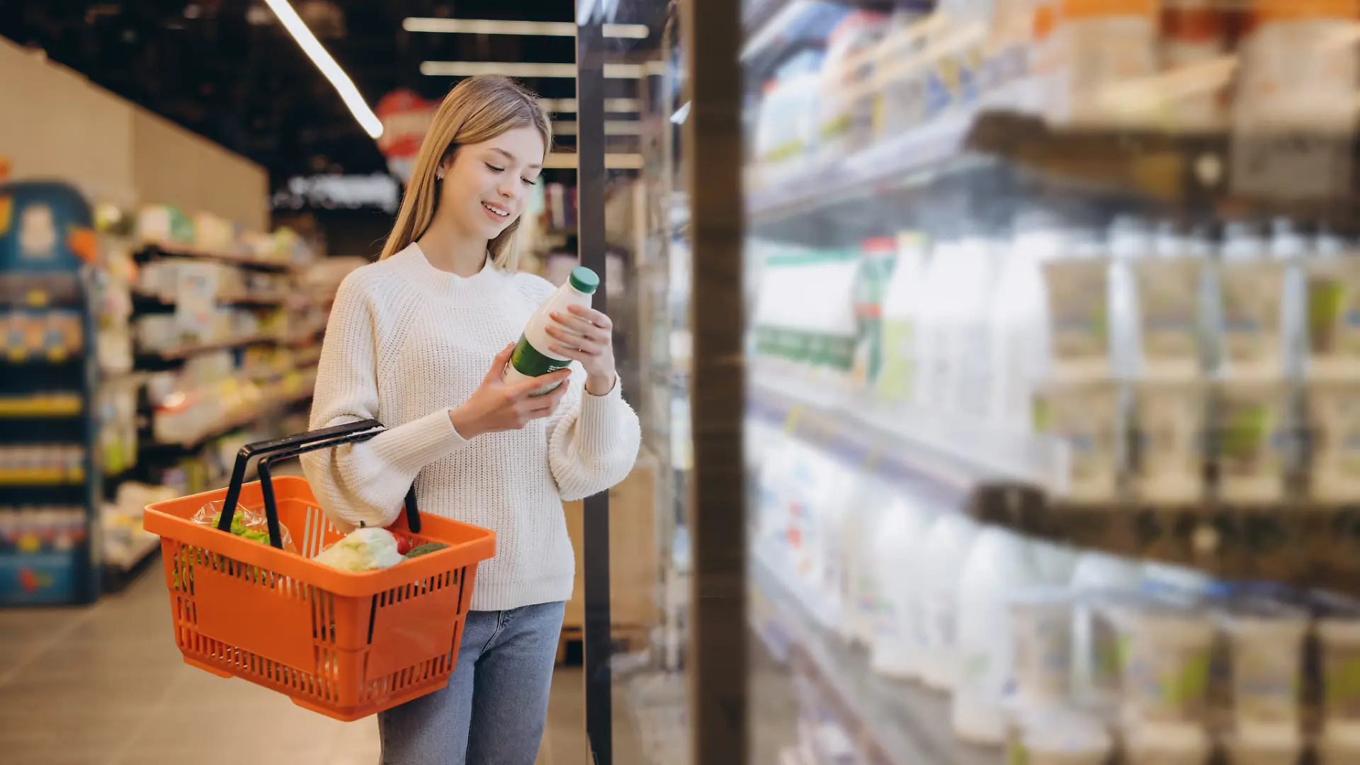 Young woman shopping groceries, holding orange basket and checking milk bottle in refrigerated section of supermarket