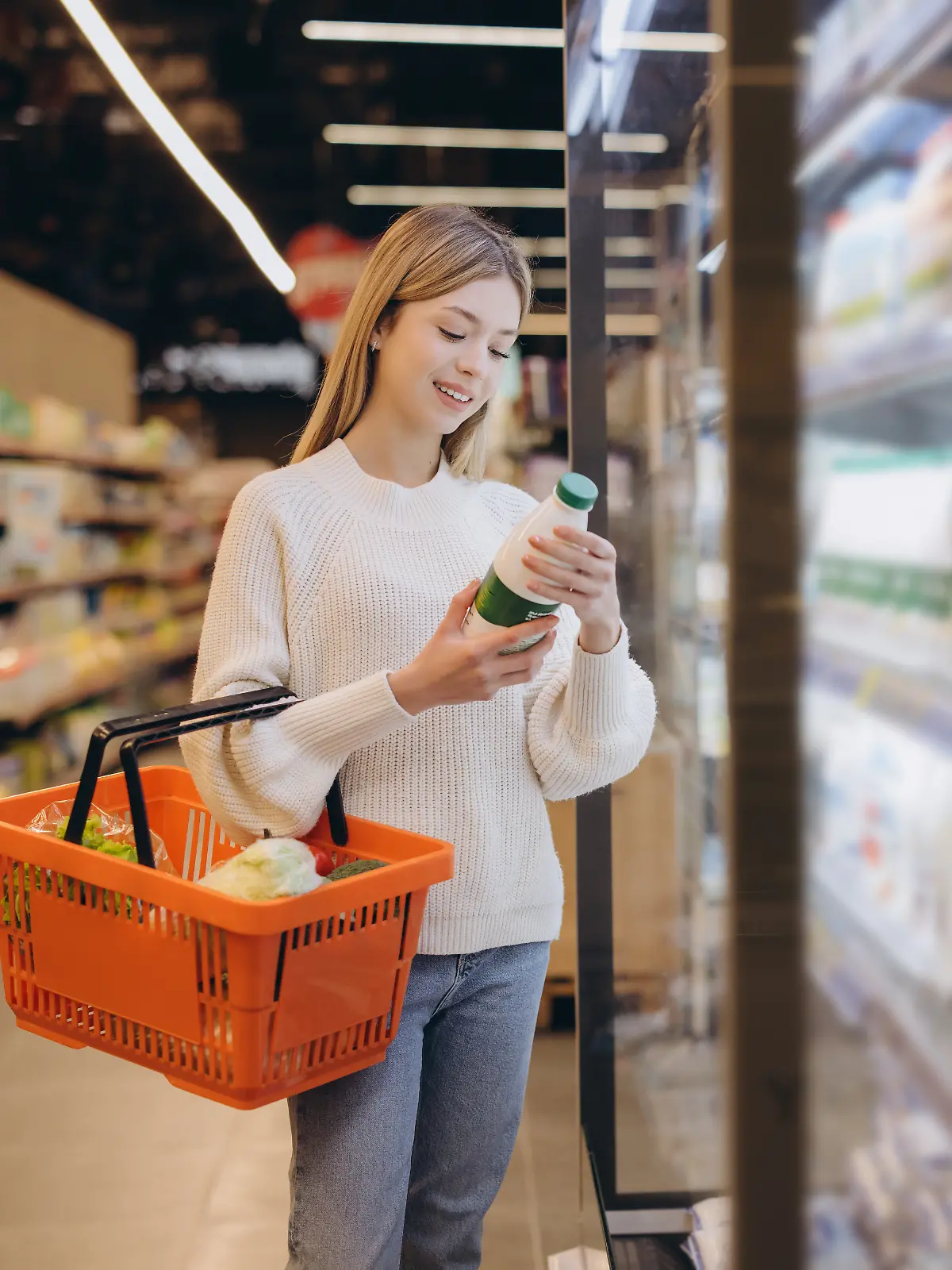 Young woman shopping groceries, holding orange basket and checking milk bottle in refrigerated section of supermarket