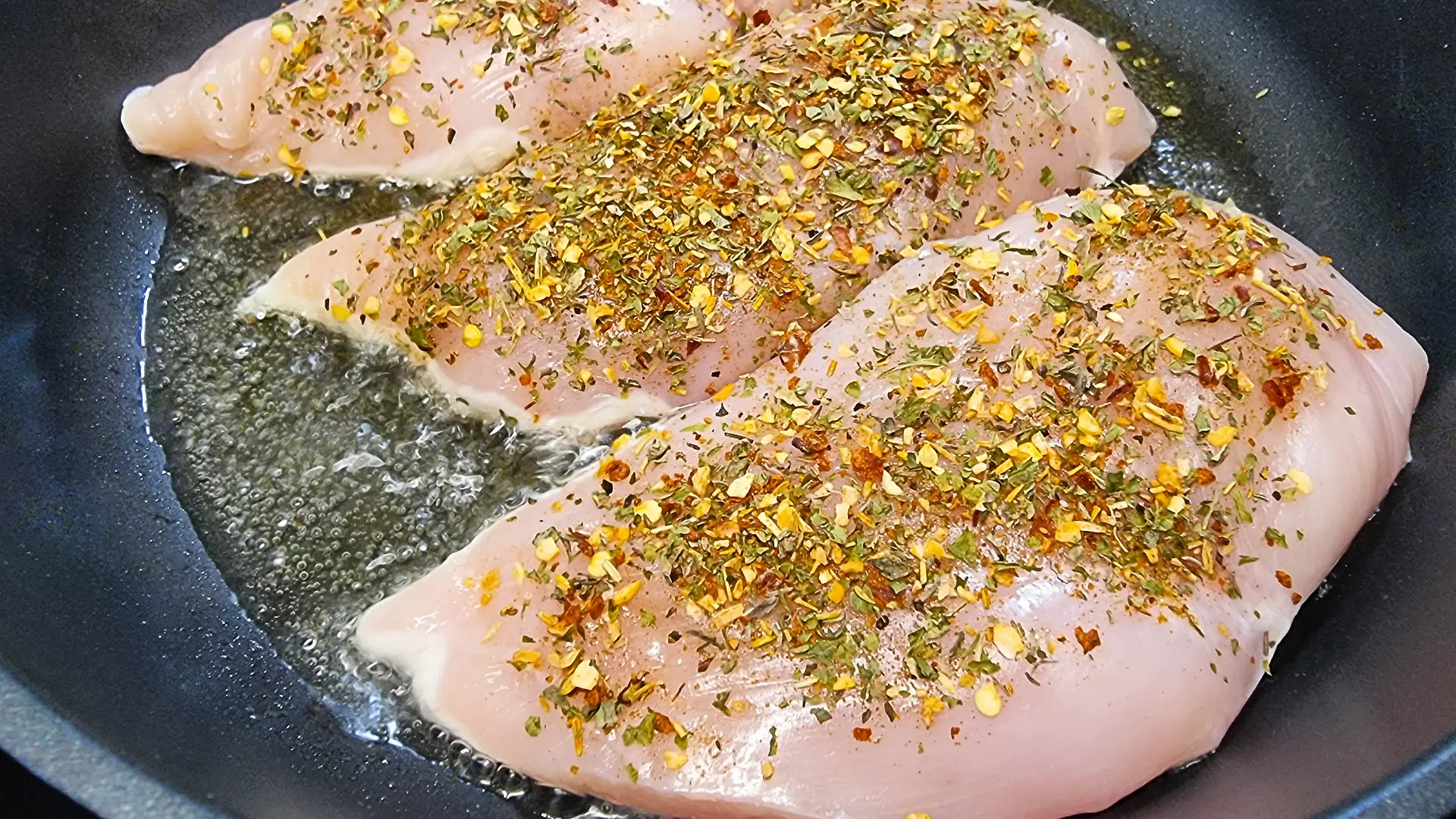 rohe Hähnchenbrust wird in der Pfanne angebraten, bestreut mit Gewürzen und Kräutern