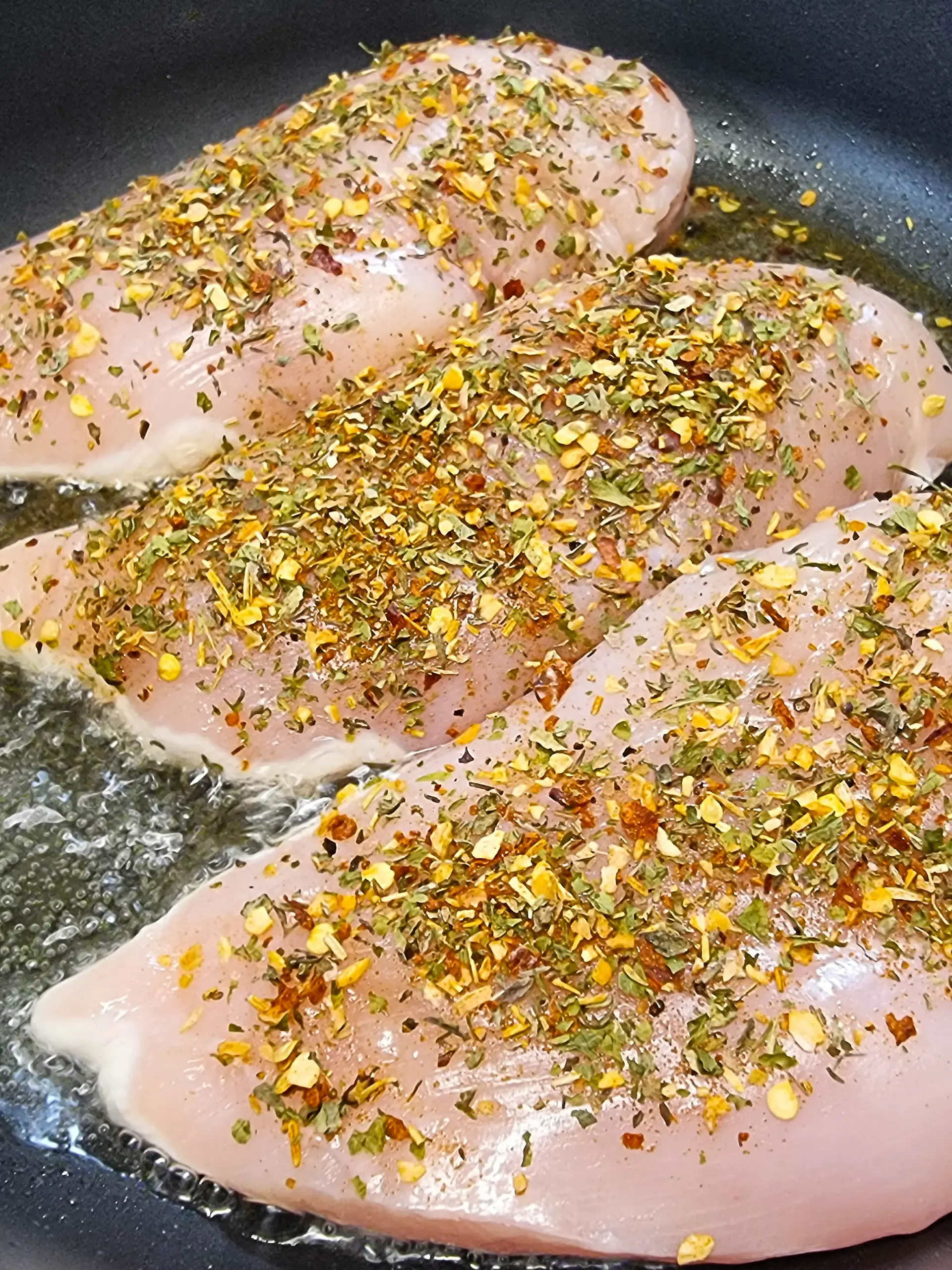 rohe Hähnchenbrust wird in der Pfanne angebraten, bestreut mit Gewürzen und Kräutern