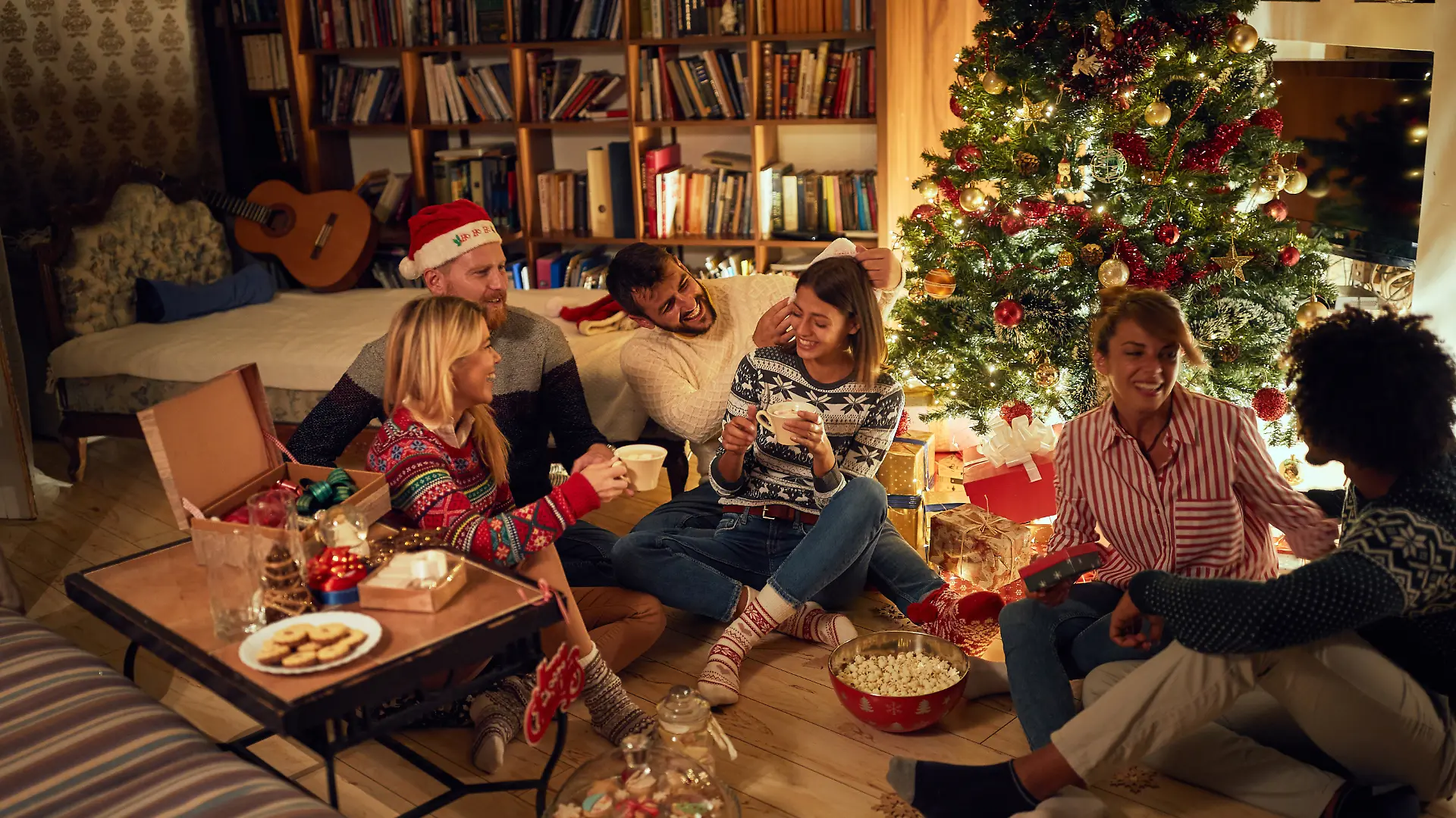 Fröhliche Freunde sitzen neben einem Weihnachtsbaum, essen Weihnachtsplätzchen, trinken Kakao und haben Spaß.